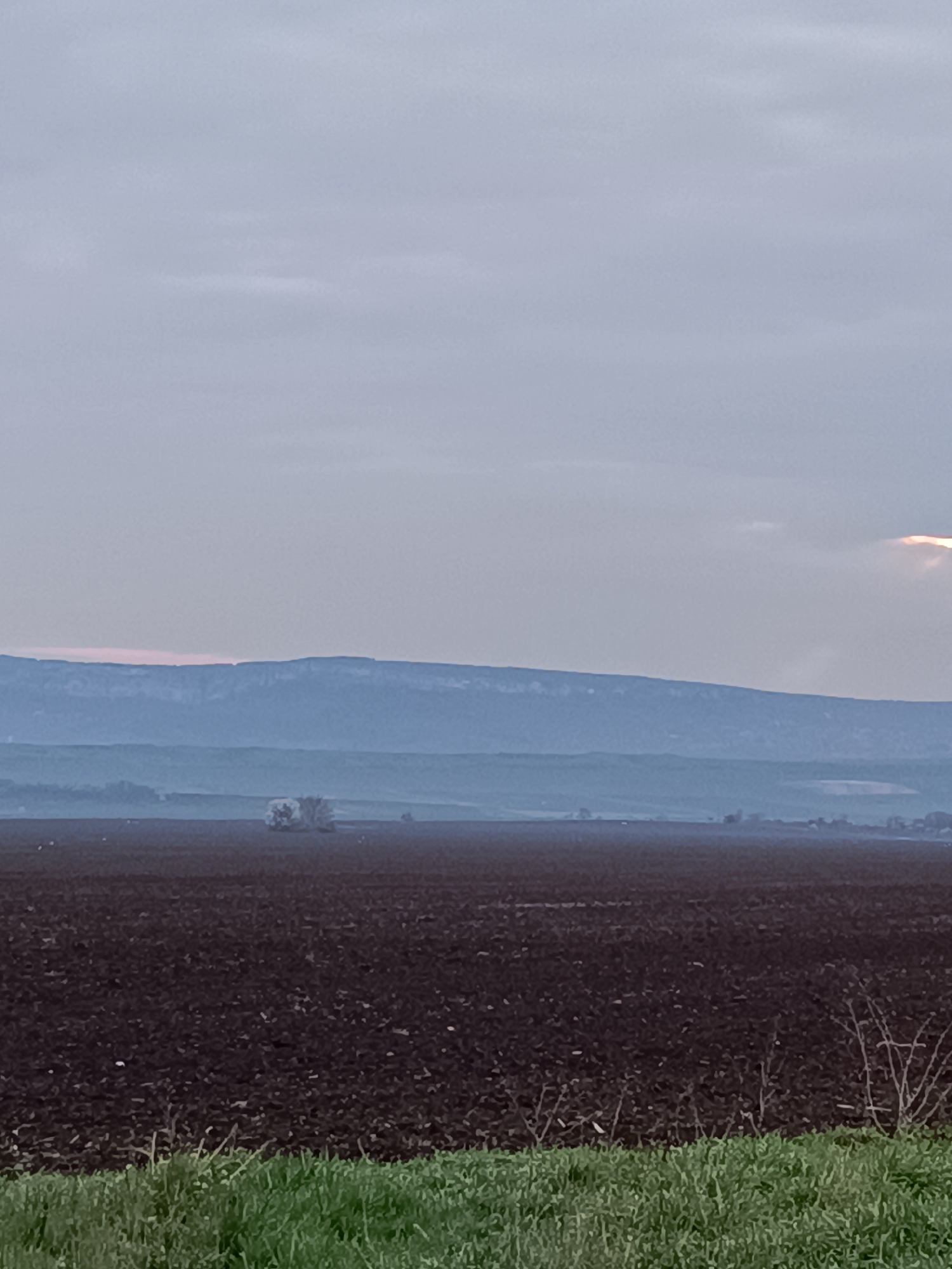 The scene depicts a rural landscape near Vasil Drumev, Bulgaria. In the foreground, green grass transitions into a field of dark, freshly tilled soil. Beyond the field, a misty haze obscures the middle distance, partially concealing scattered trees and possibly small buildings. In the background, a low mountain range is visible under a cloudy sky. A faint glow appears behind the clouds in the upper right. No people are visible in the scene.