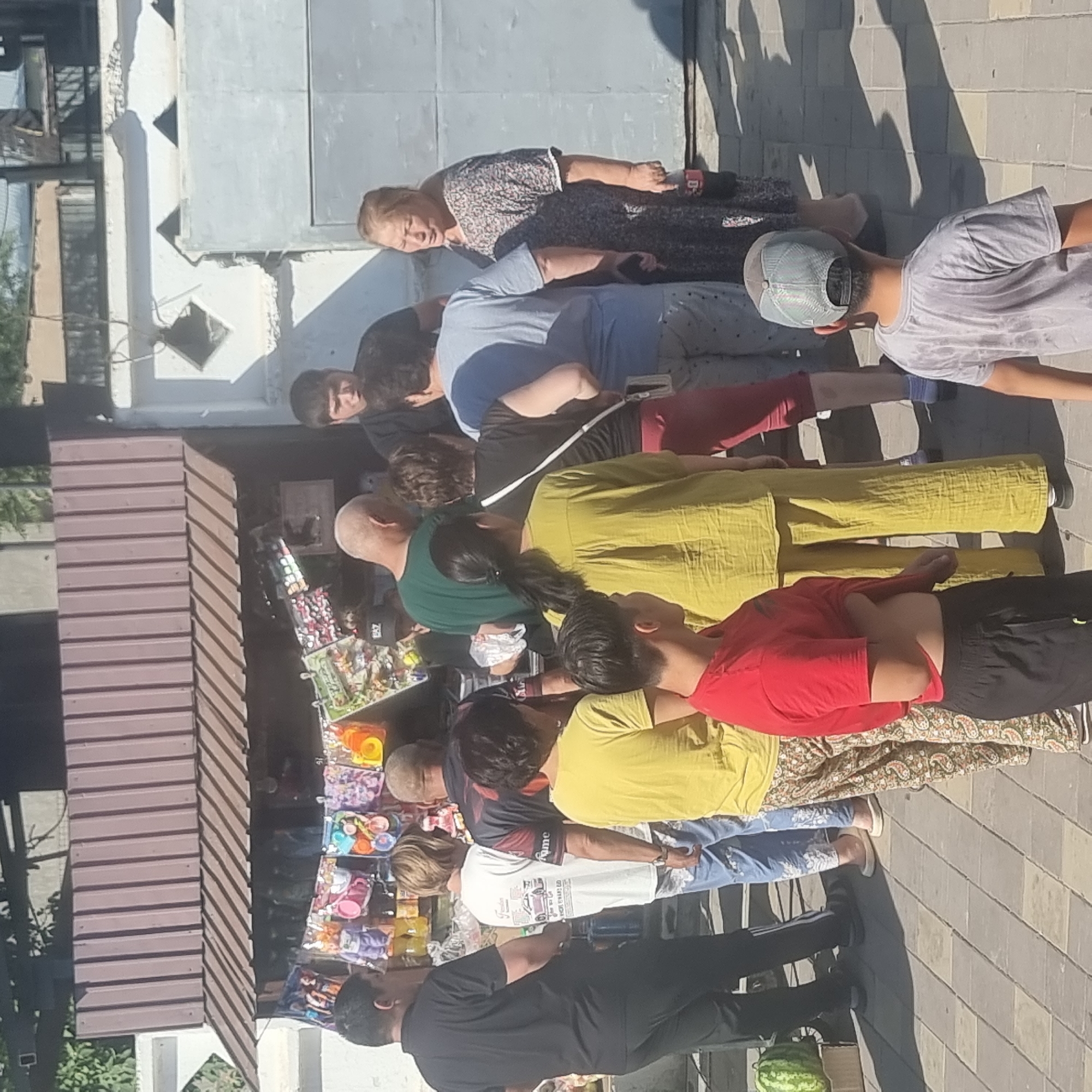 The image depicts an outdoor scene in front of a small, open-air stall in Turkistan, Kazakhstan. A group of people, appearing to be locals, are gathered around the stall. The stall displays a variety of small items, possibly toys or snacks, hanging from a rack. The people are dressed in casual clothing suitable for warm weather. Some individuals are interacting with the stall, while others are observing. The background shows a simple building structure with a corrugated metal roof and a white decorative wall. The ground is paved with interlocking bricks. The lighting suggests it is daytime, with visible shadows cast on the ground.