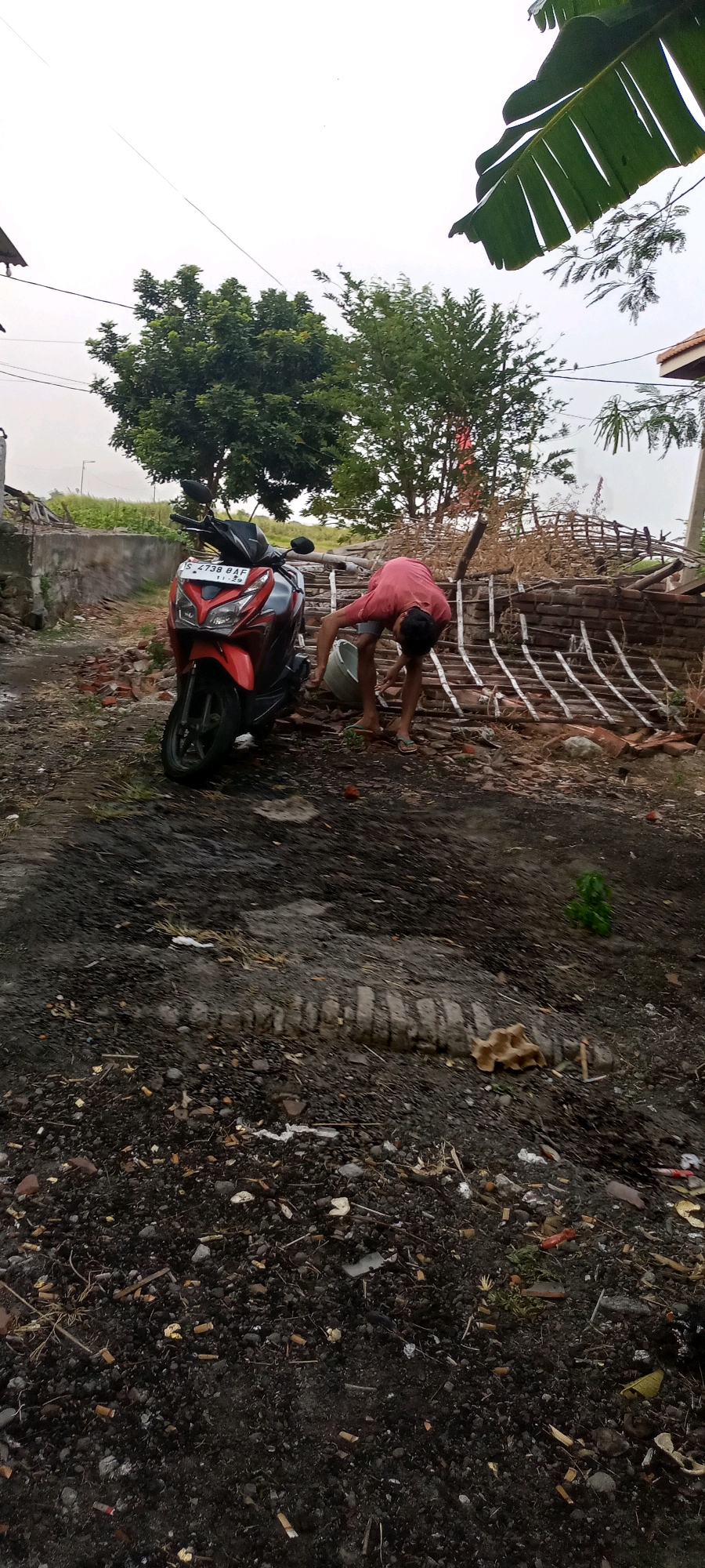 This is a medium shot, outdoor, eye-level photograph taken in Gempolklutuk, Indonesia. The scene depicts a person wearing a pink t-shirt and dark shorts, bending over to interact with a white bucket next to a red and black scooter. The scooter is parked on a dirt and debris-covered ground, with scattered bricks and wooden planks visible in the background. Trees with green foliage frame the scene from above and behind, and a cloudy sky is present. The environment appears somewhat unkempt, with the ground covered in small stones, dirt, and some refuse. A damaged brick wall structure is situated behind the scooter and the person.