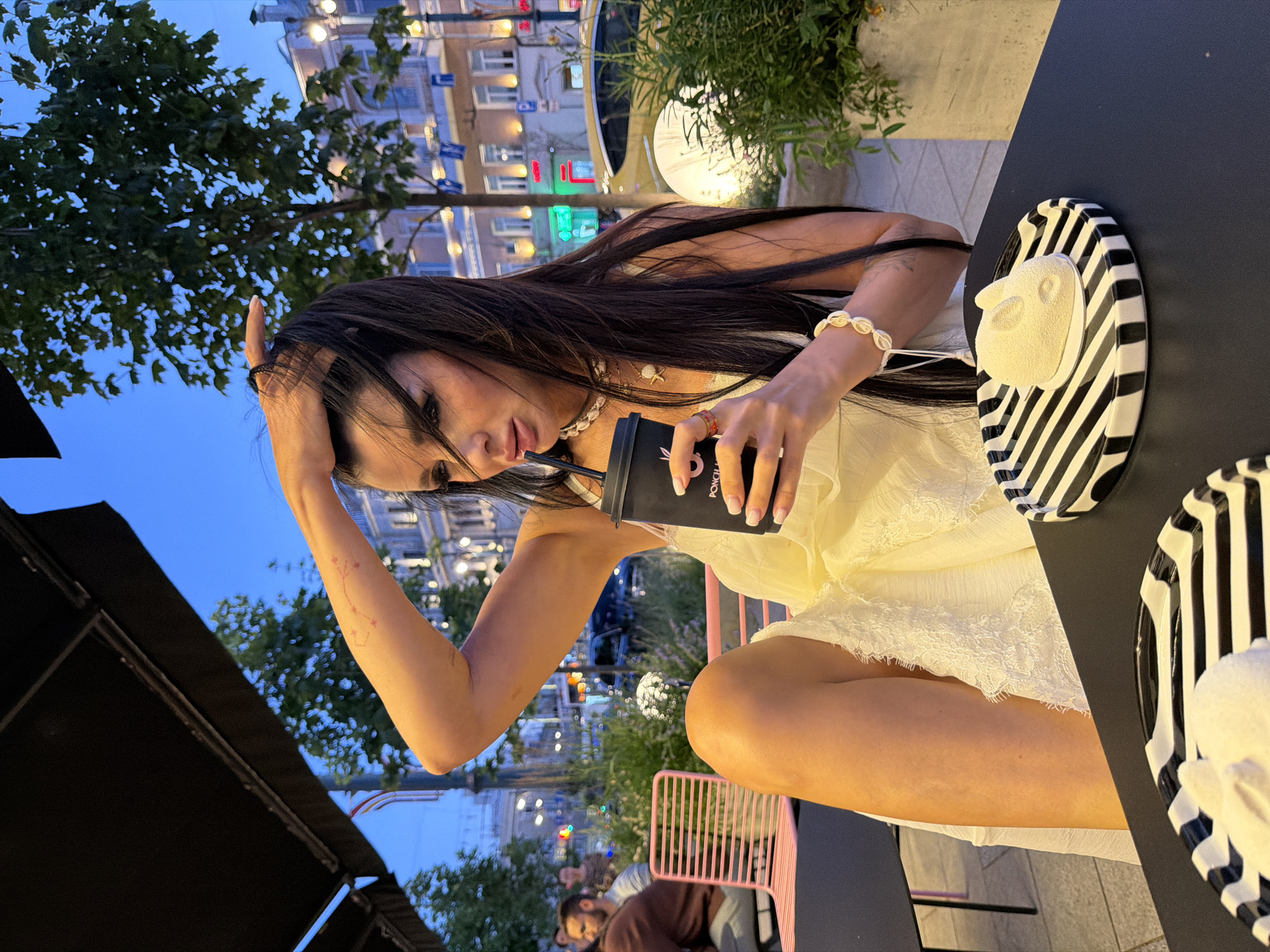 An adult female is seated at an outdoor table at twilight. She has long dark hair, is wearing a white dress, and is holding a black cup with a straw, from which she is drinking. She is also wearing jewelry, including a white beaded bracelet and a necklace. On the table in front of her is a black and white striped plate with a white, rabbit-shaped dessert. In the background, there are trees, buildings, and streetlights, suggesting an urban setting. The location is identified as Novaya Usman', Russia.