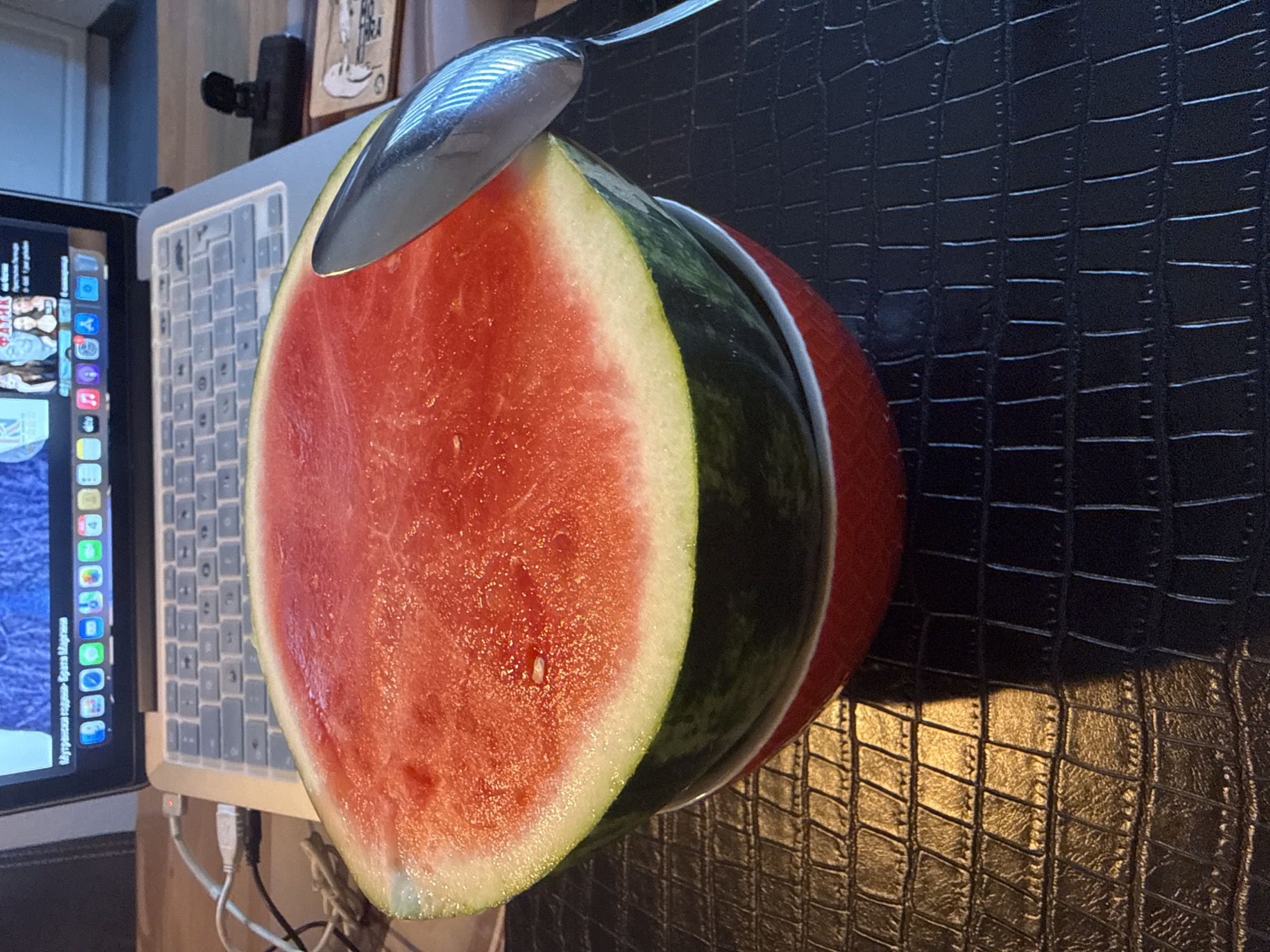 A sliced watermelon rests on a red bowl, with a spoon partially inserted into its flesh. The watermelon is positioned in front of an open laptop. The scene is set on a dark, textured surface, possibly a table. The background shows a blurred view of the laptop screen and keyboard. The location is identified as Marum, Netherlands.
