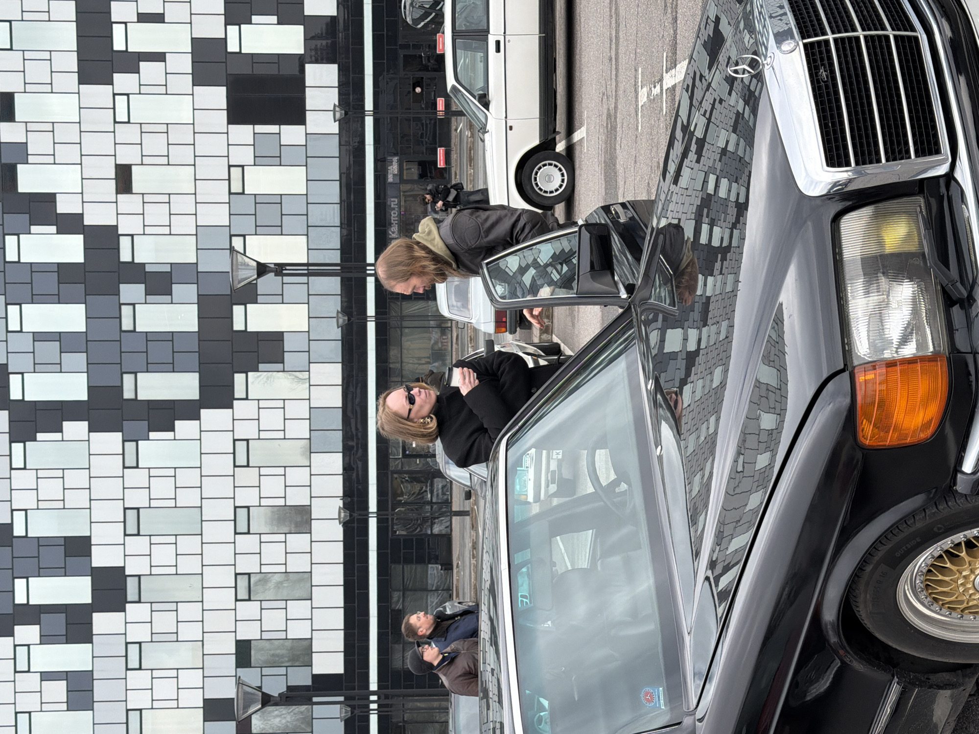 The image shows a black Mercedes-Benz parked on a street in Saint Petersburg, Russia. Two people are standing near the open driver's side door of the car. One person, a woman with short blonde hair and sunglasses, is standing inside the car. Another person, a man with long hair, is leaning into the car. Another man in a brown jacket is near the driver. The background features a modern building with a grid-like facade and a white car is parked behind the Mercedes. The building’s facade reflects the street and people. The image is taken from a low angle, focusing on the car and the people nearby.