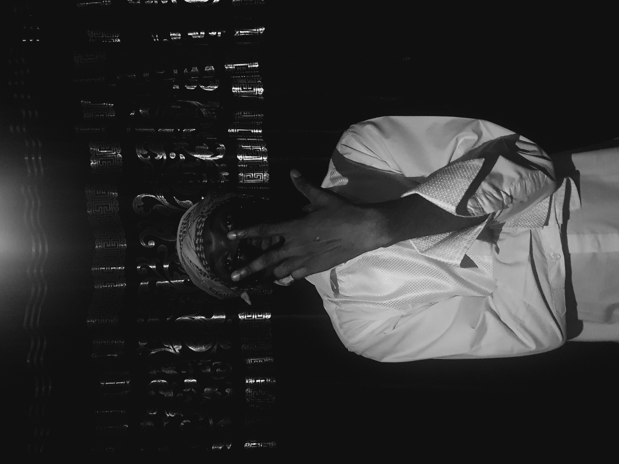 A black and white photograph depicts a person in a white shirt and bandana, positioned to the right side of the frame. The person is making a hand gesture. The background is dark and reflects light, seemingly a decorated surface. The location is indeterminable within Lokoja, Nigeria, without further evidence.