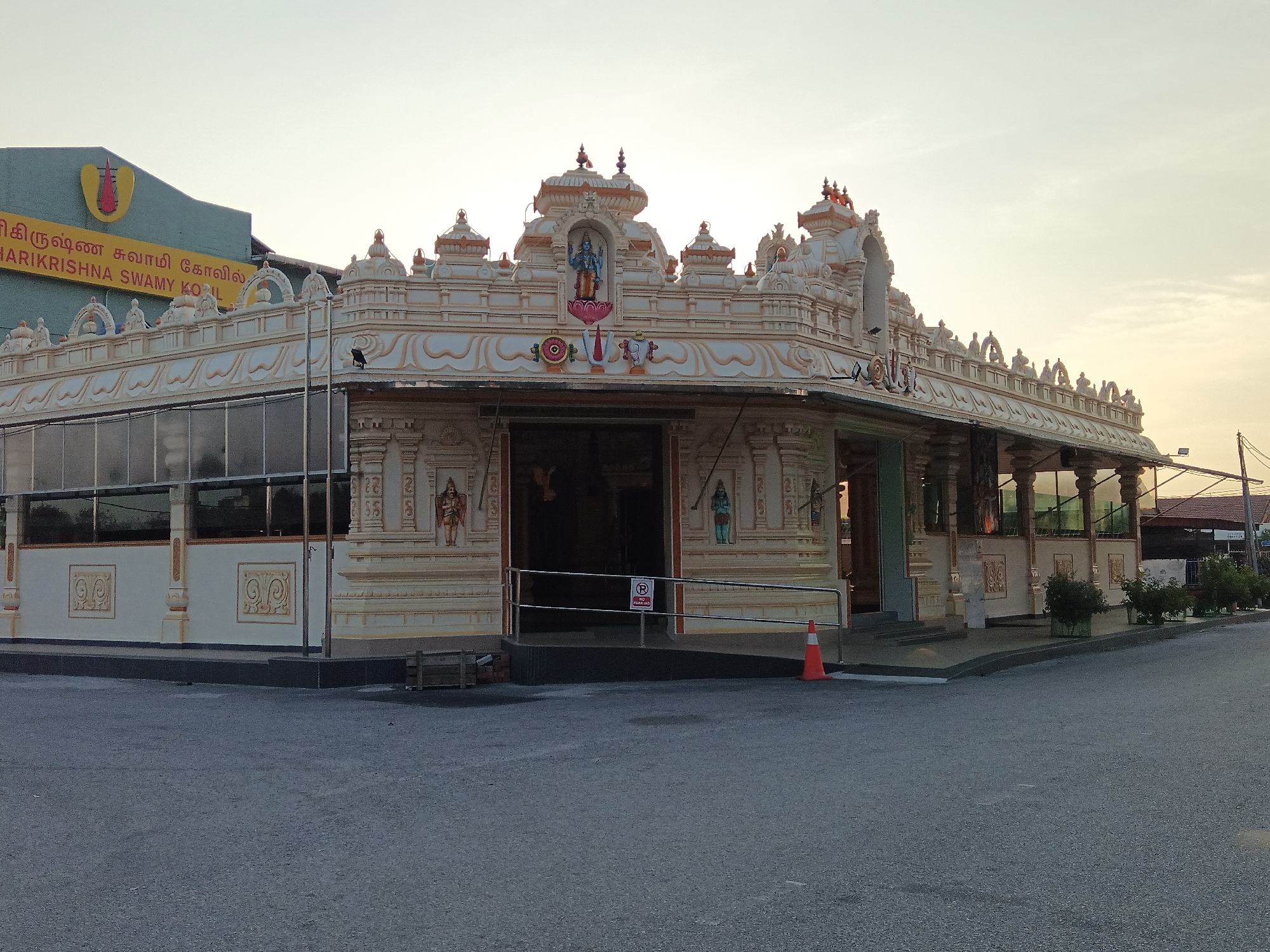 A low-angle, medium shot captures the exterior of the Sri Krishnar Swamy Koil temple in Klang City, Malaysia. The temple, adorned with intricate carvings and statues, is primarily white and orange. A yellow sign with Tamil script and a red flame symbol is visible on a teal building in the background. In the foreground, an asphalt pavement surrounds the temple. An orange traffic cone stands near the temple entrance, and a "No Parking" sign is displayed on a railing. The sky is a pale yellow, suggesting it is either dawn or dusk. The scene is devoid of any people.