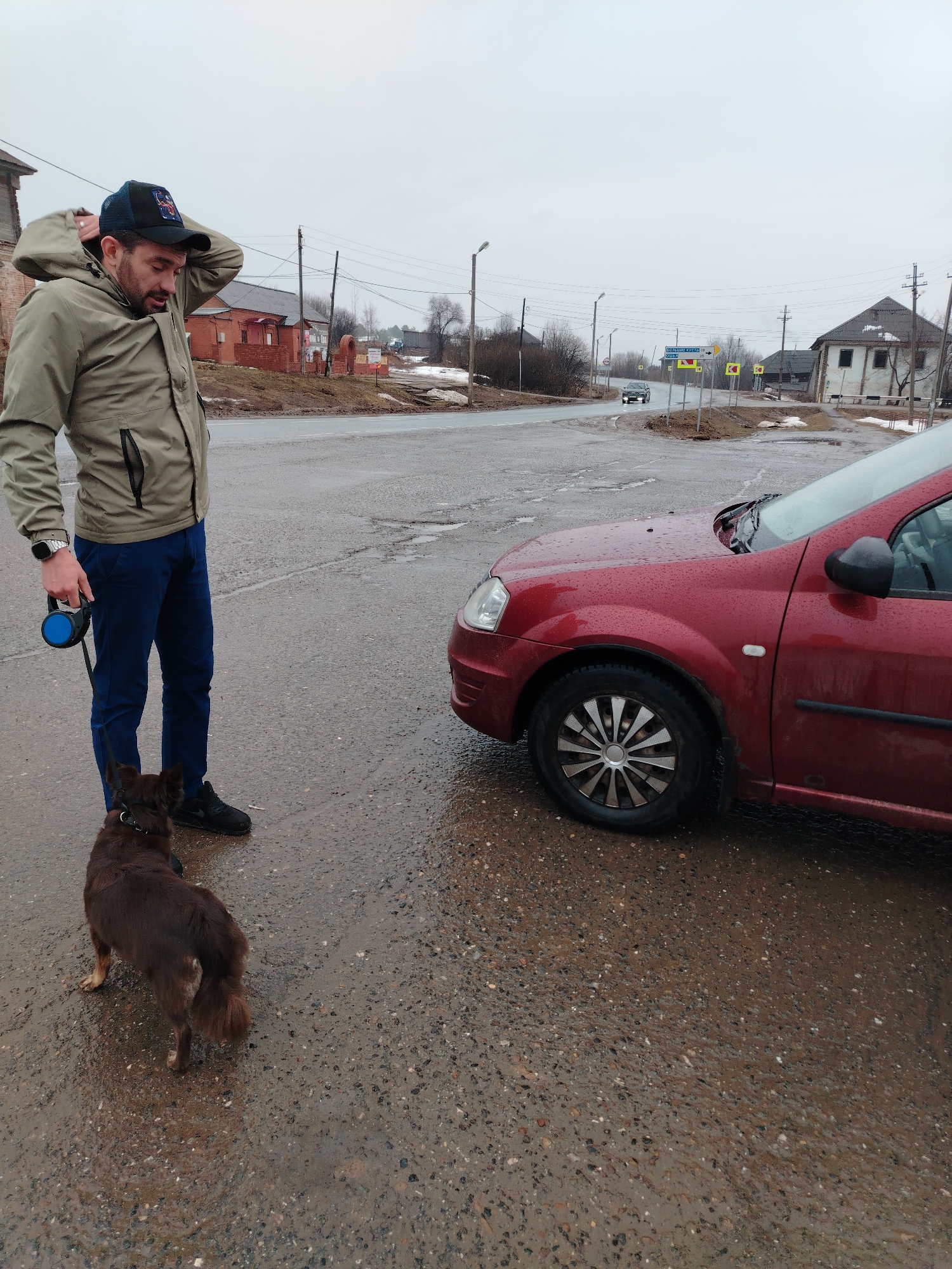 A medium shot captures a man and a dog standing on a wet asphalt surface near a red car. The man, dressed in a light green jacket, blue pants, and black shoes, is adjusting his baseball cap while holding a blue retractable leash. A medium-sized brown dog is positioned to his left.
The car, partially visible on the right, is red with water droplets on its surface. The background reveals a typical street scene in the city of Большая Уса, Russia, characterized by modest buildings, utility poles, and road signs. Patches of melting snow are visible. Another vehicle is on the road in the distance. The weather appears overcast.