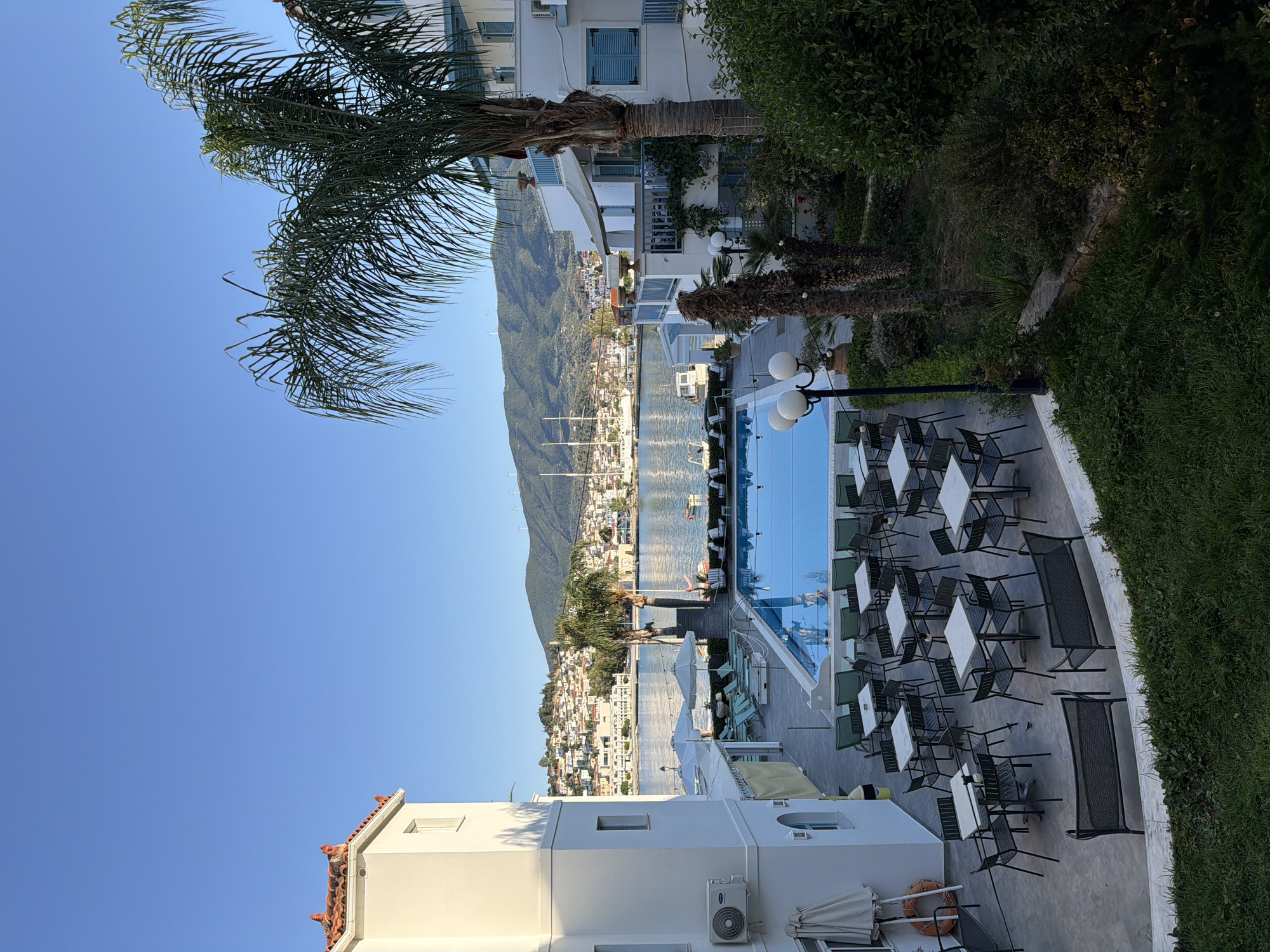 This vibrant image captures a scenic coastal view, likely in Poros, Greece, depicting a resort or hotel area overlooking a bay. The scene is devoid of people, creating a peaceful ambiance.
In the immediate foreground, an outdoor dining or lounge area is visible, furnished with numerous white rectangular tables and dark-colored chairs, some with slatted designs. To the left of this seating arrangement, a bright blue swimming pool reflects the clear sky. Several multi-globe street lamps illuminate the area. Lush green hedges and grass frame the patio space, which is bordered by white building facades. A large palm tree extends its fronds upwards on the left, while smaller palms dot the landscape. An air conditioning unit, bearing the "CARRIER" brand, is mounted on one of the white buildings, which also features a folded white umbrella and a visible lifebuoy on a wall.
Beyond the foreground amenities, a serene body of water stretches across the midground, dotted with several boats, including sailboats with tall masts. On the opposite shore, a picturesque town unfolds, characterized by numerous white buildings with distinct red-tiled roofs, climbing up a verdant hillside. In the distant background, majestic green mountains dominate the horizon, with several wind turbines discernible along their ridge. The sky is a clear, bright blue, suggesting a sunny day, likely late morning or early afternoon given the quality of light and shadows. The overall impression is one of a tranquil and beautiful Mediterranean vacation destination.