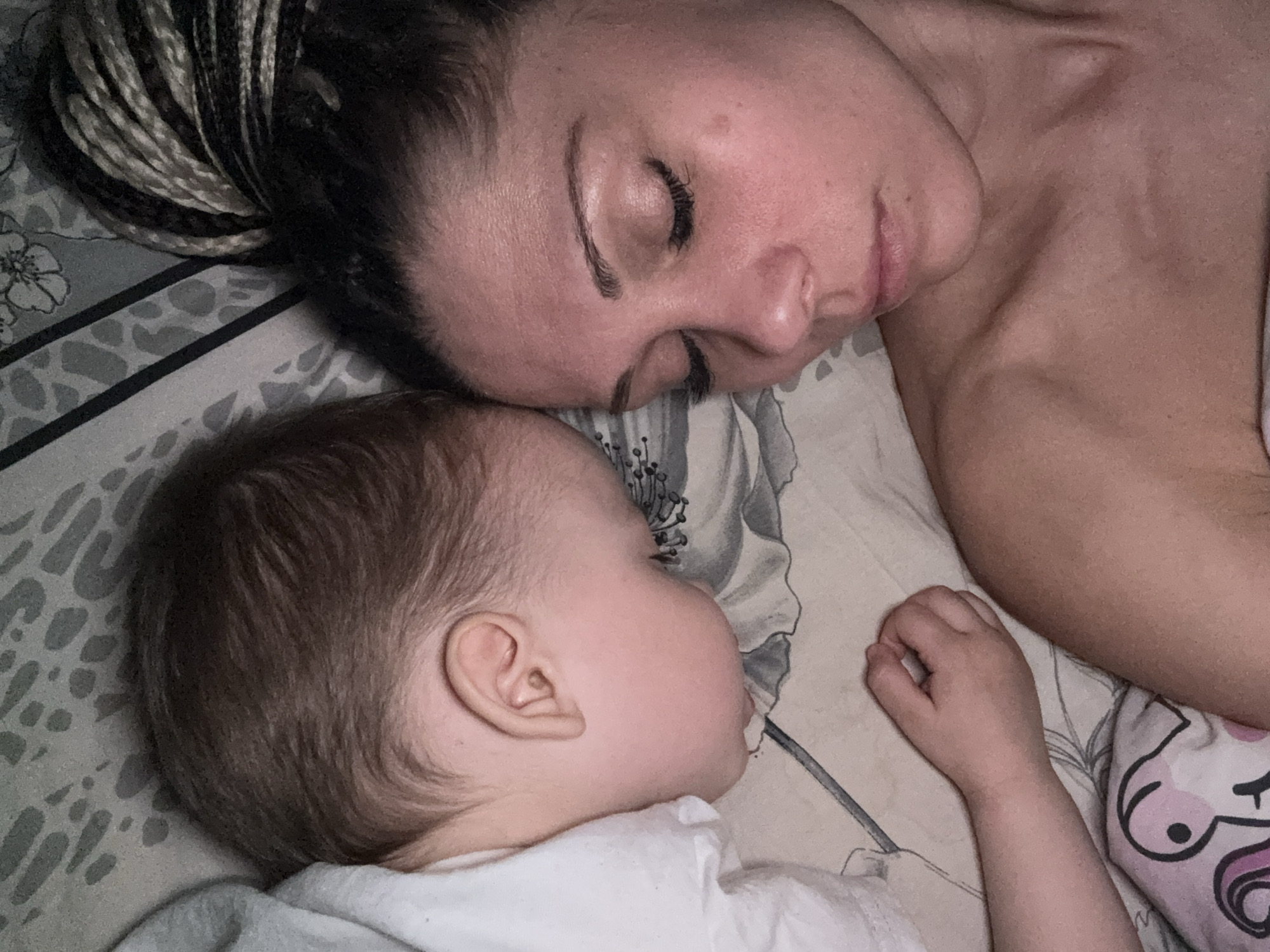 The image depicts a woman and a baby sleeping side-by-side on a bed. The woman has her eyes closed and appears to be resting peacefully. She has a fair complexion and her hair is styled in braids. The baby is lying close to the woman, also with eyes closed, suggesting sleep. The baby has light skin and short brown hair. They are lying on a bed with patterned sheets. Based solely on the visual information available, no connection to the city of Rusanovo, Russia can be established.