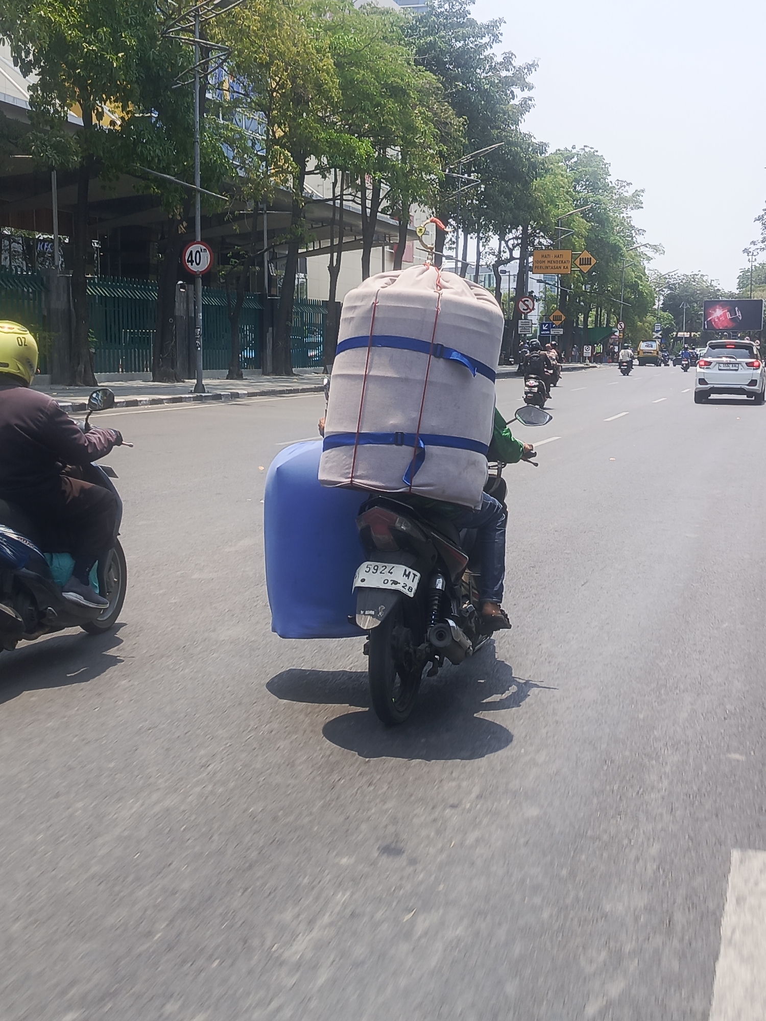 A motorcycle rider with a large cylindrical load secured to the back navigates a city street. The load, covered in a beige fabric and strapped with blue bands, is significantly larger than the motorcycle itself. A blue tarp is visible on the side of the motorcycle, possibly part of the load. The rider, wearing a dark long-sleeved shirt and blue jeans, has a yellow helmet. The motorcycle's license plate reads "5924 MT". In the background, other vehicles, including cars and motorcycles, are present on the road. Trees line the street, and buildings are visible behind them. Traffic signs are also present, including one indicating a speed limit of 40 km/h and another with yellow diamond shapes. The scene suggests a busy urban environment in Surabaya, Indonesia, likely on a sunny day given the bright lighting and shadows.