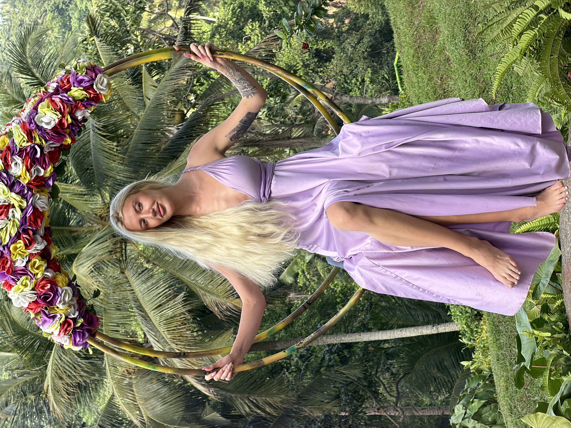 In Abiansemal, Indonesia, a woman with long blonde hair and tattoos on her arm sits on a swing decorated with colorful flowers. She is wearing a flowing lavender dress. The swing is set against a backdrop of lush green palm trees and foliage, suggesting a tropical outdoor location.