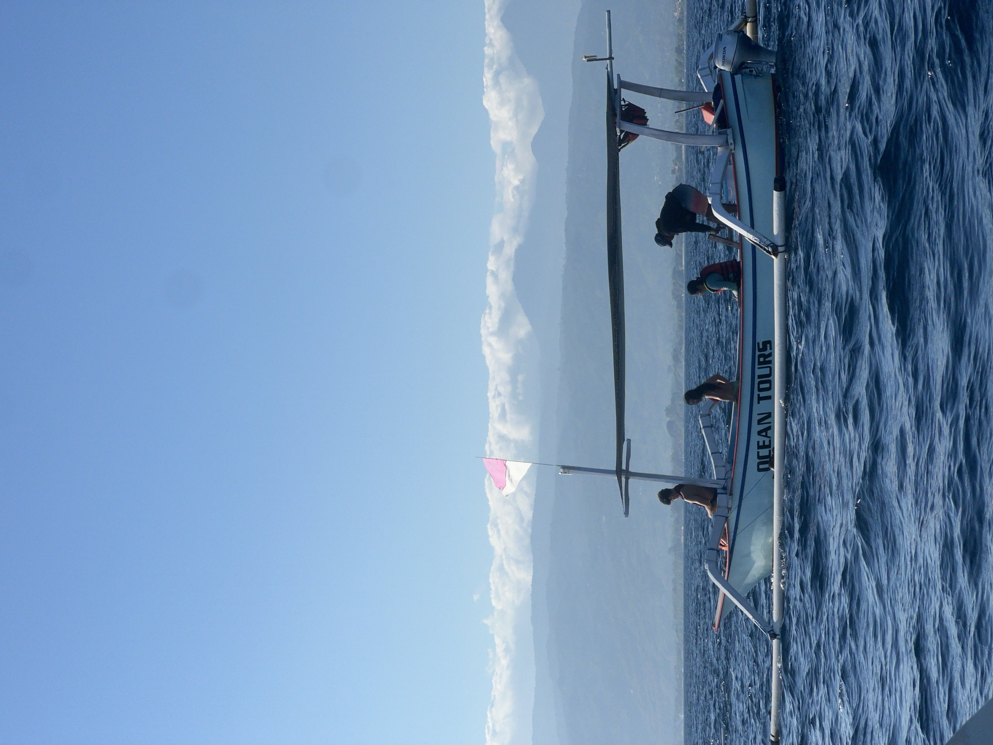A small, white motorboat with outriggers is at sea under a clear blue sky. The boat has a canopy providing shade for some of its occupants. The words "OCEAN TOURS" are visible in dark letters on the side of the boat. There are at least four people on board. One person, wearing a dark jacket and shorts, is leaning over the edge of the boat. Another person, wearing a life vest, is seated. Two other individuals, appearing to be young adults, are also seated on the boat, one of whom is shirtless. A pink and white flag is attached to a pole on the boat. In the background, mountains are visible, partially obscured by clouds, and a shoreline with trees suggests land. The water is a deep blue with moderate waves. The scene is likely coastal, potentially near Seririt, Indonesia, given the boat's design which is common in Indonesian waters for tourism and transport.