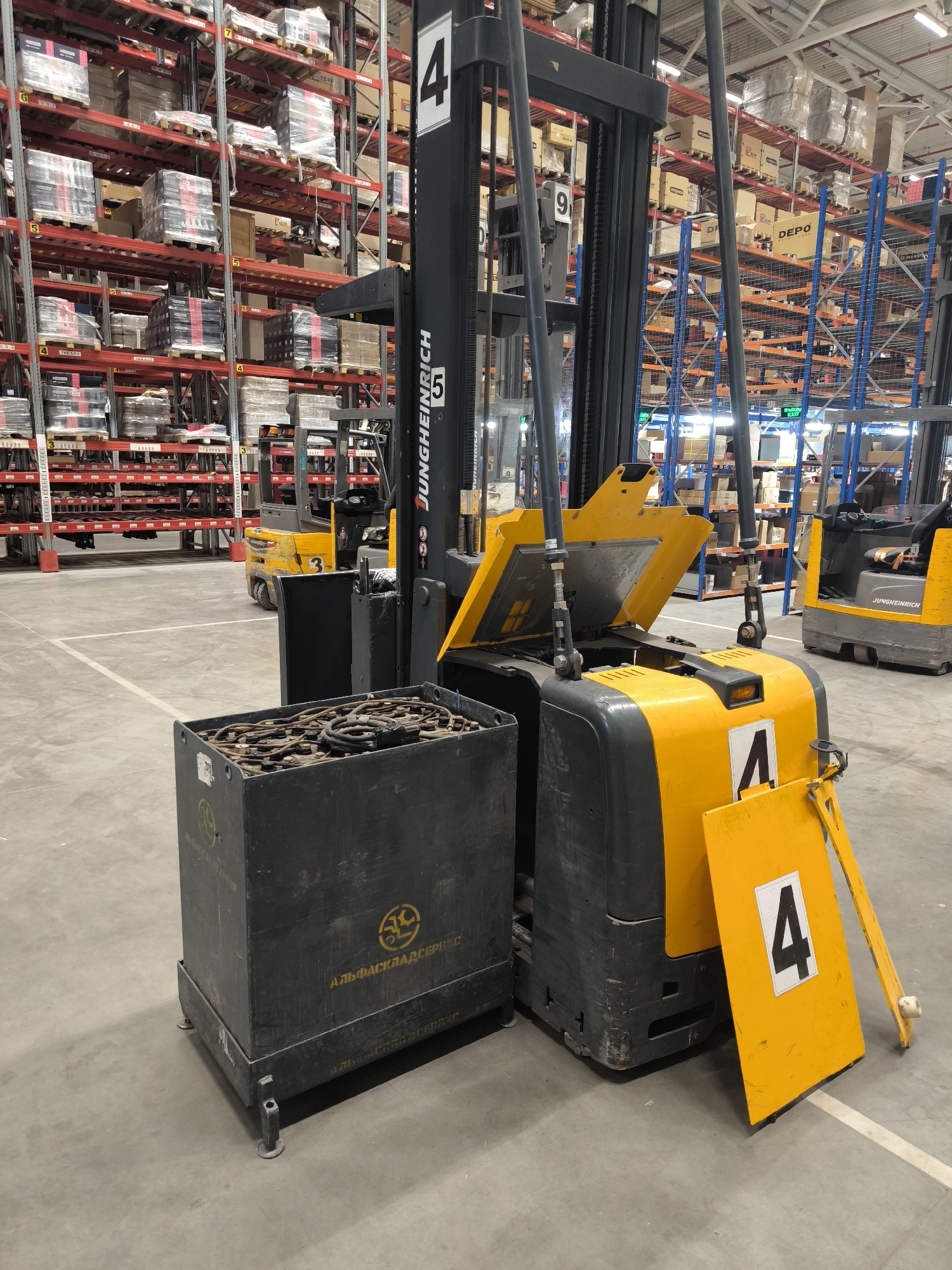 The image shows the interior of a warehouse in Domodedovo, Russia, likely an industrial or logistics facility. The warehouse is filled with tall shelving units stacked with various boxed goods, indicating organized storage. In the foreground, a yellow and black Jungheinrich forklift is prominently featured. It is partially obscured by a black metal container filled with what appears to be used forklift batteries, connected by thick cables. A yellow safety sign with the number "4" in a white square is attached to the side of the forklift. Another yellow forklift, also bearing the "Jungheinrich" brand, is visible in the background to the right. The floor is concrete and marked with white lines, suggesting a designated pathway or area. The overall environment appears to be a functional, active warehouse space. There are no people visible in the frame.