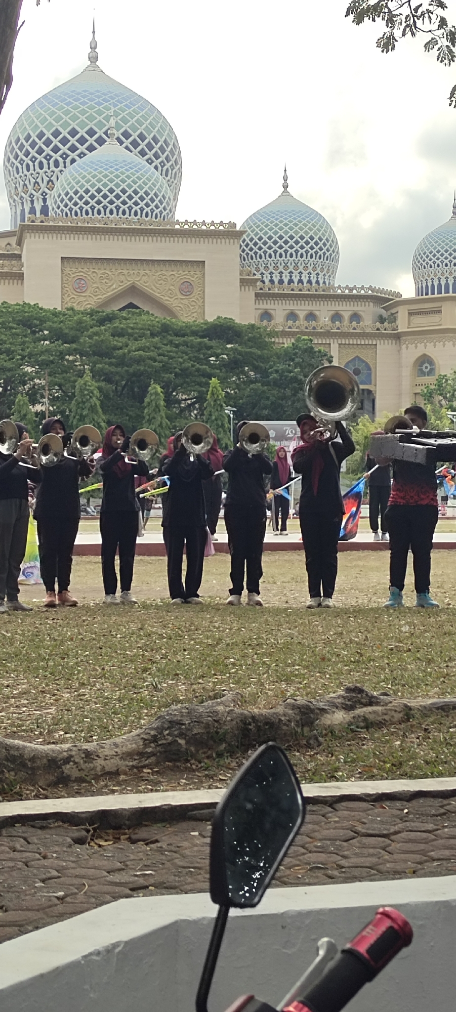 A marching band, composed of at least eight individuals wearing black uniforms and headscarves, stands in a line on a grassy field. They are holding brass instruments, including tubas and trumpets, and appear to be performing or practicing. In the background, a large, ornate building with multiple domes, possibly a mosque, is visible, set against a cloudy sky. Trees and foliage are also present in the background. The scene suggests an outdoor event or practice session taking place in a city that is likely Lhokseumawe, Indonesia, given the architectural style and the commonality of such events in educational or cultural settings. The lower foreground of the image shows a motorcycle's rearview mirror and handlebars, indicating the photograph was taken from a vehicle.