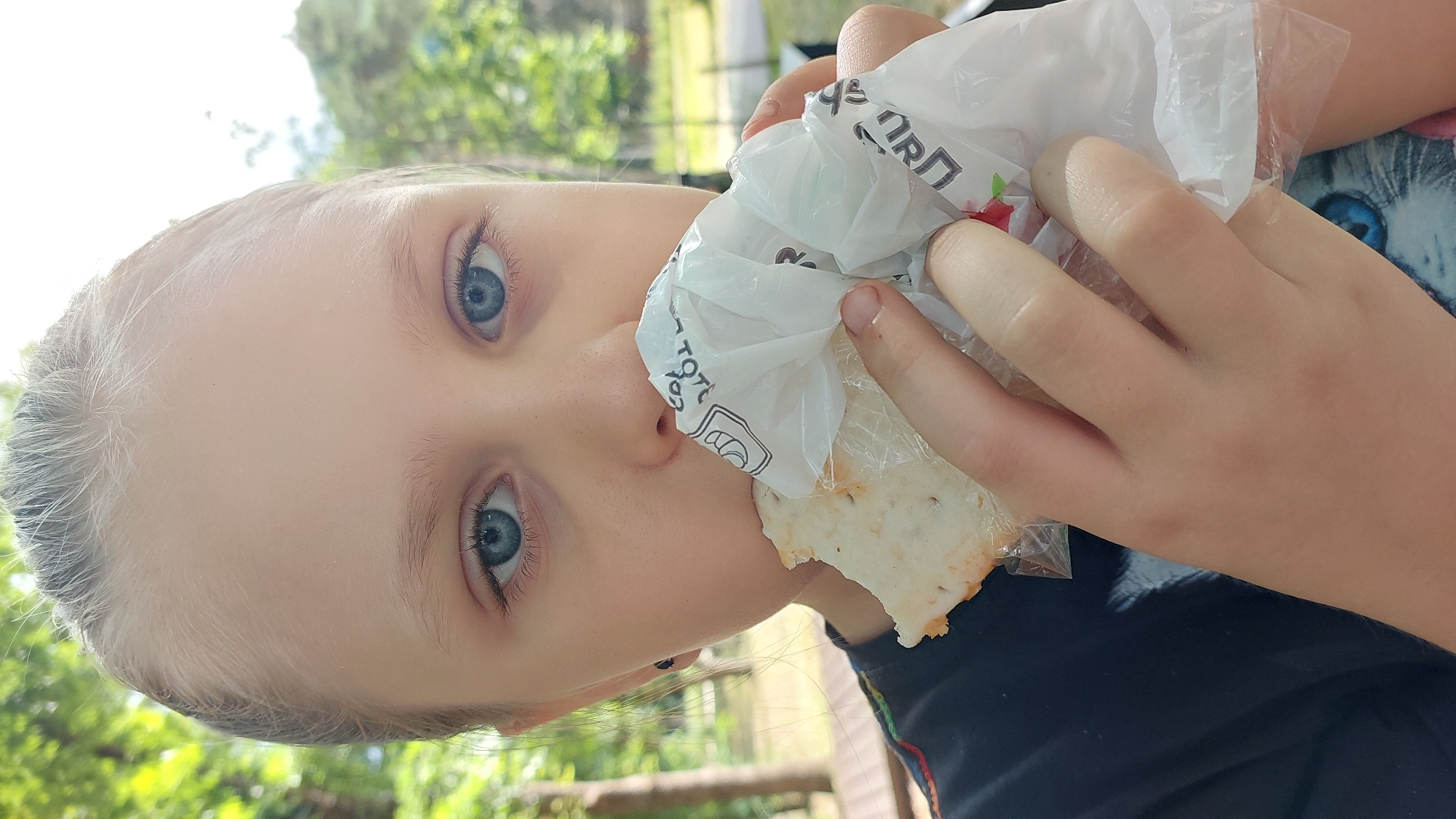 A young person with light skin and blonde hair is shown in a close-up, sideways shot as they eat. Their large blue eyes are wide open and looking directly at the camera. They are holding a white paper wrapper, possibly from a food item, up to their mouth with both hands, biting into something wrapped inside. The wrapper appears to have some black text and a red graphic on it. The background is blurred and shows green foliage and trees, suggesting an outdoor setting. The lighting is bright, indicating it is daytime, likely sunny. The person's facial expression appears neutral or perhaps slightly curious. The clothing visible is a dark shirt with a colorful trim on the sleeve. The overall impression is a candid moment of someone enjoying a snack outdoors.
