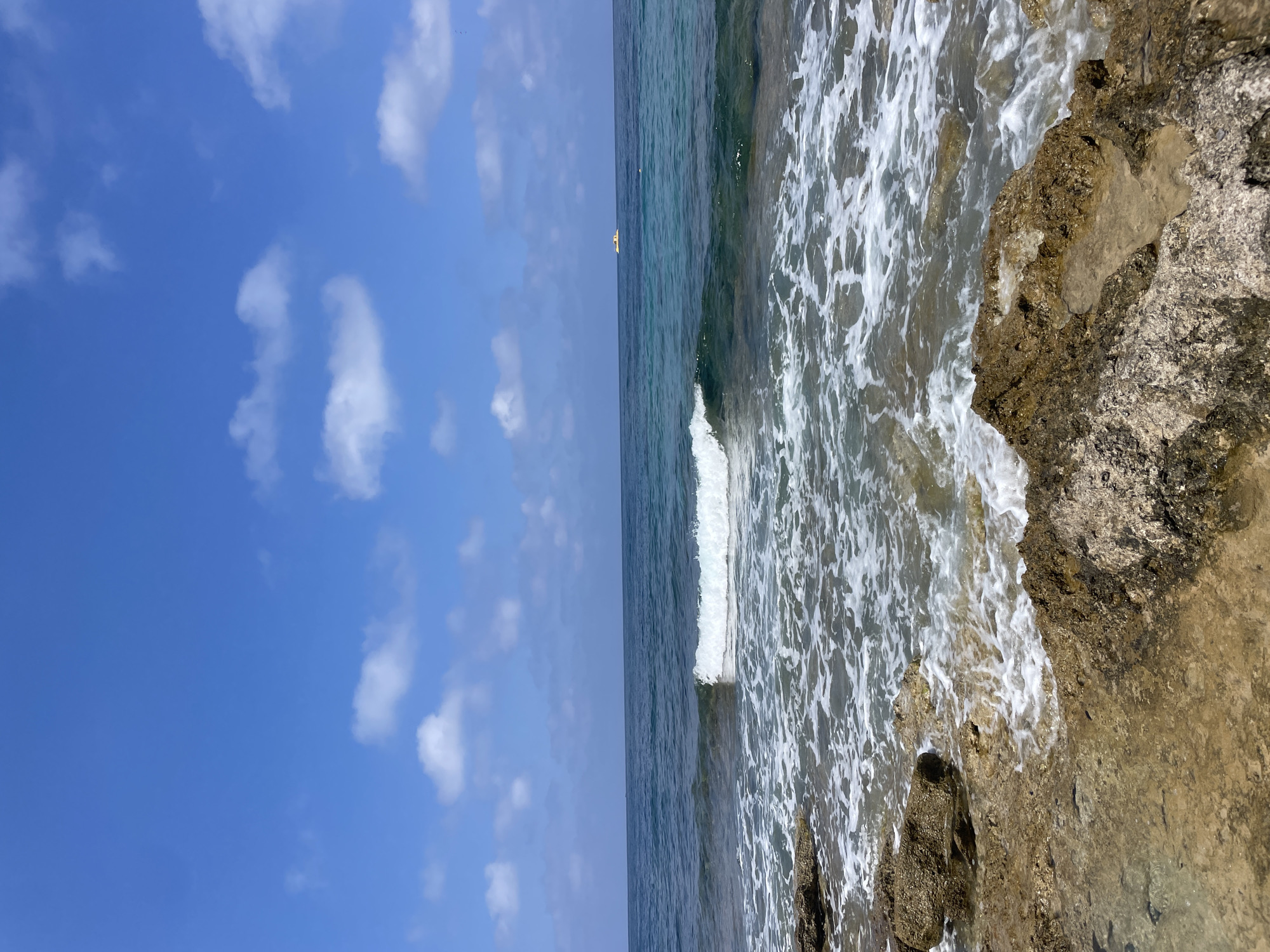 The image depicts a coastal scene in Paphos, Cyprus, under a partly cloudy blue sky. The foreground is dominated by rough, wet rocks of various brown and gray hues, with some showing a yellowish tint from sand or mineral deposits. Waves are breaking against these rocks, creating white foam and ripples in the shallow water. The sea extends into the mid-ground, transitioning from a clear, shallow greenish-blue near the shore to a deeper, darker blue further out. A single, small, yellow object, possibly a boat or a buoy, is visible on the horizon in the distance. The water is calm with gentle waves. The environment is natural and serene, with no people or animals visible. The lighting suggests it is daytime, likely mid-morning or afternoon, with the sun casting a bright light.