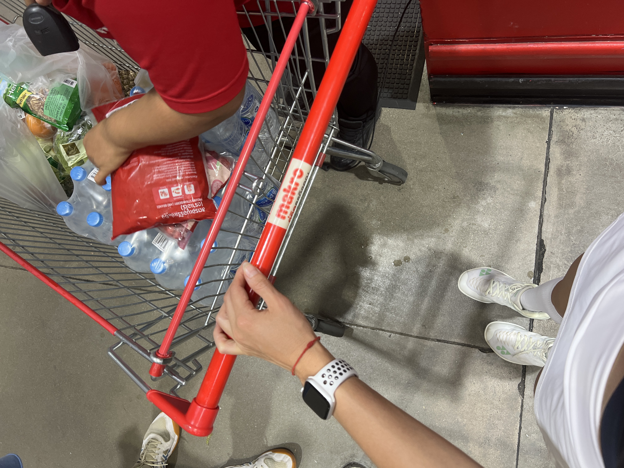 Here is a forensic description of the image:
The image captures an eye-level perspective of the inside of what appears to be a Makro store in an unspecified city. The primary focus is on a red Makro shopping cart filled with groceries, including bottled water, frozen strawberries (Aro Brand), fresh produce, and other packaged goods. The cart is being handled by two individuals, as indicated by their hands and arms visible in the frame. One person wears a red shirt, and their hand is reaching into the cart. The other person, wearing an Apple Watch with a white band, is holding the cart's handle. The floor is concrete, and the legs and feet of two people are visible: one wearing white sneakers and the other wearing beige sneakers. A red checkout counter is visible in the background. The overall impression is that the people are shopping in a Makro store and filling a cart with groceries.