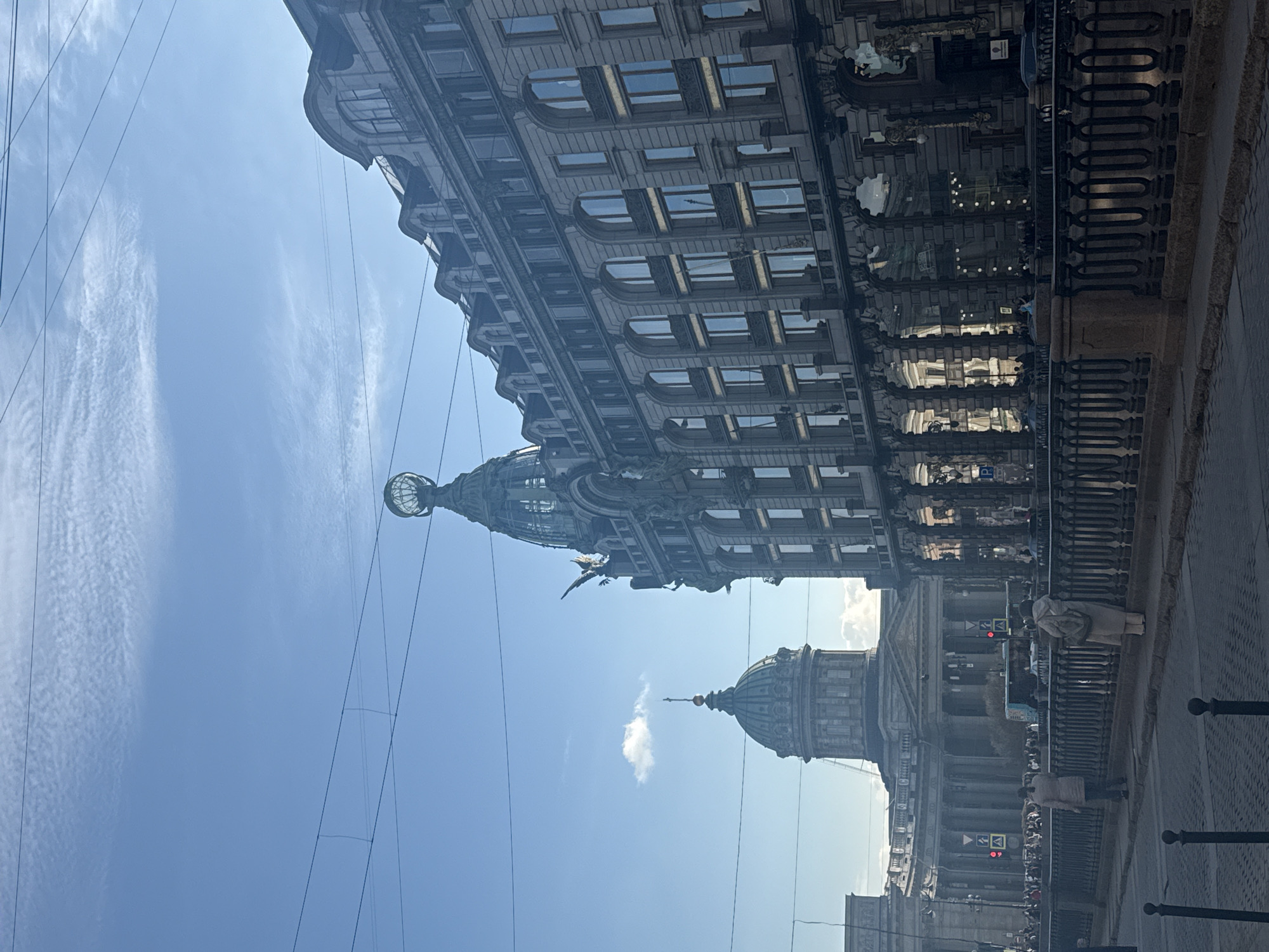 The image shows a daytime view in Saint Petersburg, Russia. The Singer House (Dom Knigi) is seen on the left, characterized by its globe-topped dome and elaborate architecture. In the background, the dome of the Kazan Cathedral is visible. The scene includes overhead power lines. Pedestrians can be seen along a street or embankment with a wrought-iron fence. Street signs and traffic lights are present. The sky is blue with scattered clouds.