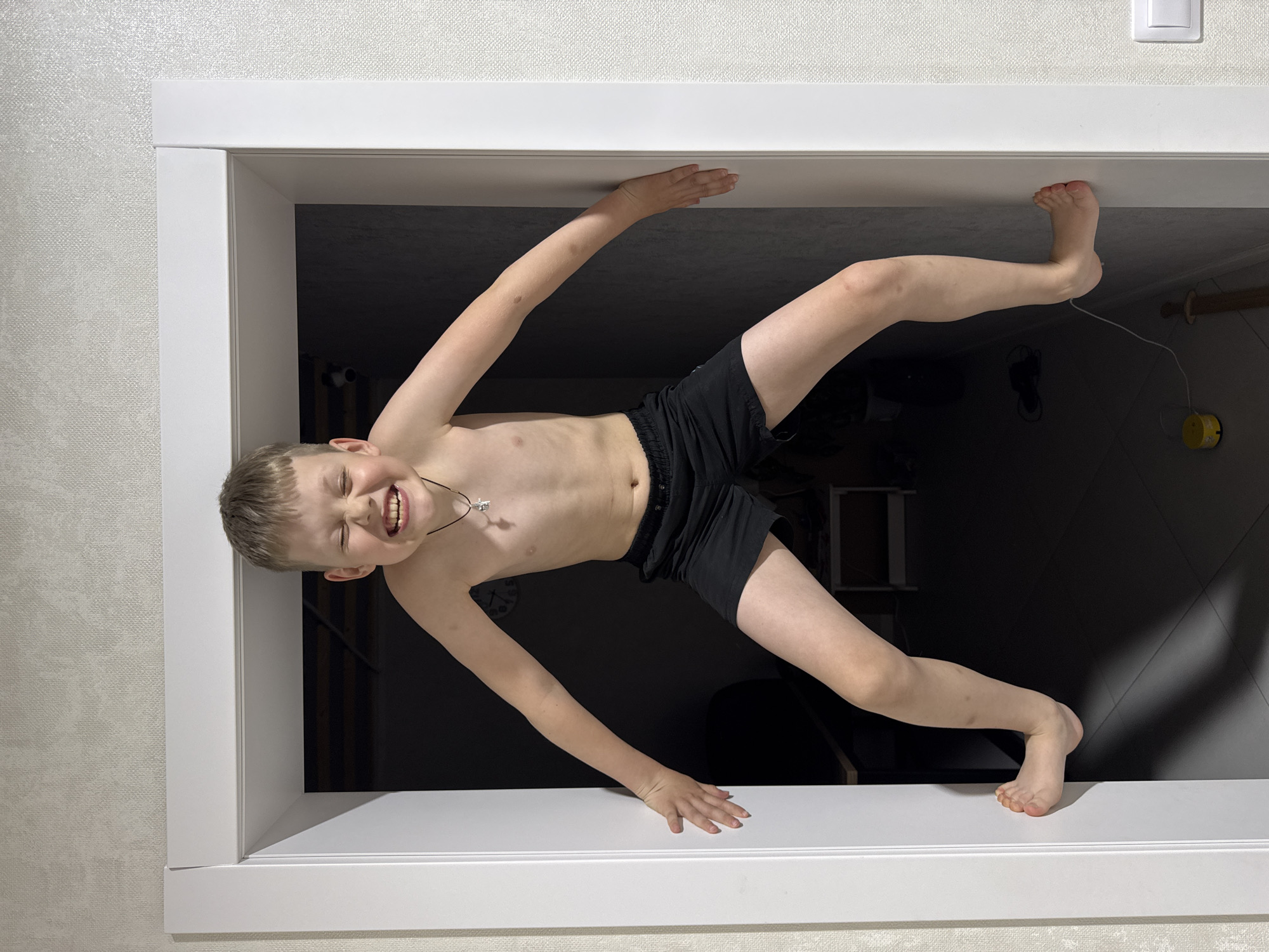 A young boy, appearing to be between 5 and 10 years old, is depicted in a playful, acrobatic pose, suspended horizontally within a white interior door frame. He is shirtless and wears black shorts, with a silver cross pendant visible around his neck. The boy's face is illuminated, showing a wide, joyful smile with his eyes squinted, indicating laughter or intense enjoyment from his activity.
He uses his hands and feet to brace himself against the upper and lower sections of the door frame, showcasing flexibility and strength. The background directly behind him, viewed through the doorway, is dark and indistinct, with faint glimpses of what might be furniture or room features, including a small yellow object on the floor to the right. The immediate foreground shows a light-colored wall with a subtle, textured wallpaper pattern. The door frame itself is white and modern. The indoor setting is presumably a residence in Gornyi (Dvurechenskoe S/P), Russia. The time of day is not clearly indicated, but the dark area beyond the doorway might suggest a dimly lit room or nighttime. No text is visible in the image.
