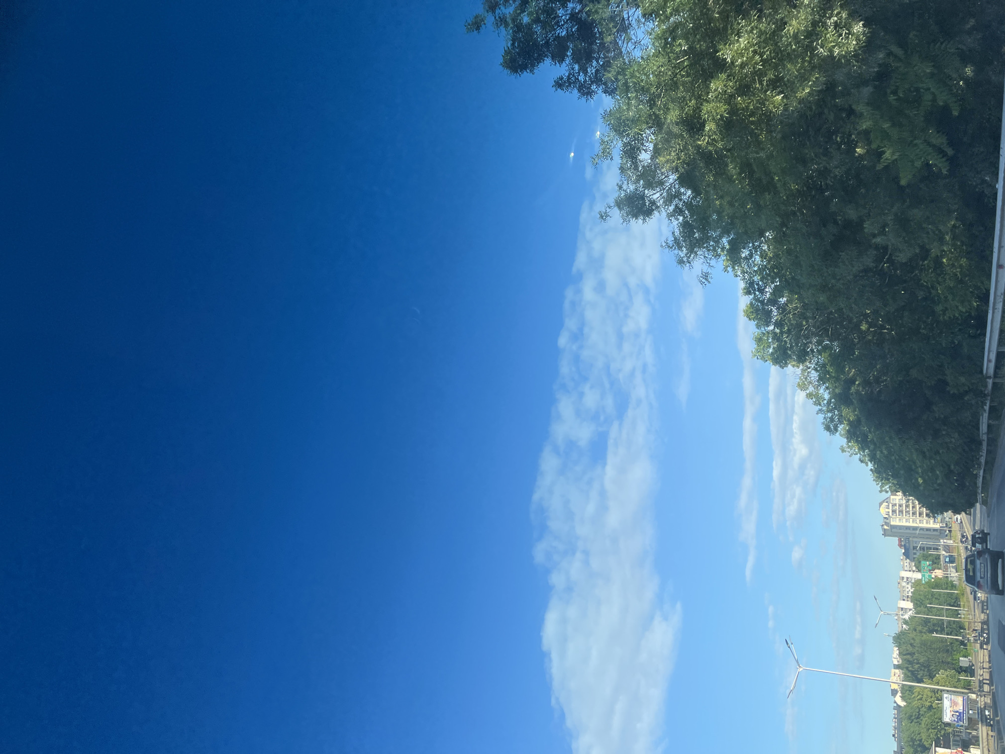 The image depicts a view from a vehicle traveling on a highway. The sky is a clear, vibrant blue with scattered white clouds. On the right side of the frame, a dense line of lush green trees lines the highway. In the distance, city buildings are visible, with some appearing to be residential or office towers. The highway itself has multiple lanes, and several cars are driving on it, including a dark gray sedan in the foreground. Streetlights are also present along the side of the road. The overall scene suggests a daytime view of a city fringe or suburban area during a clear day.