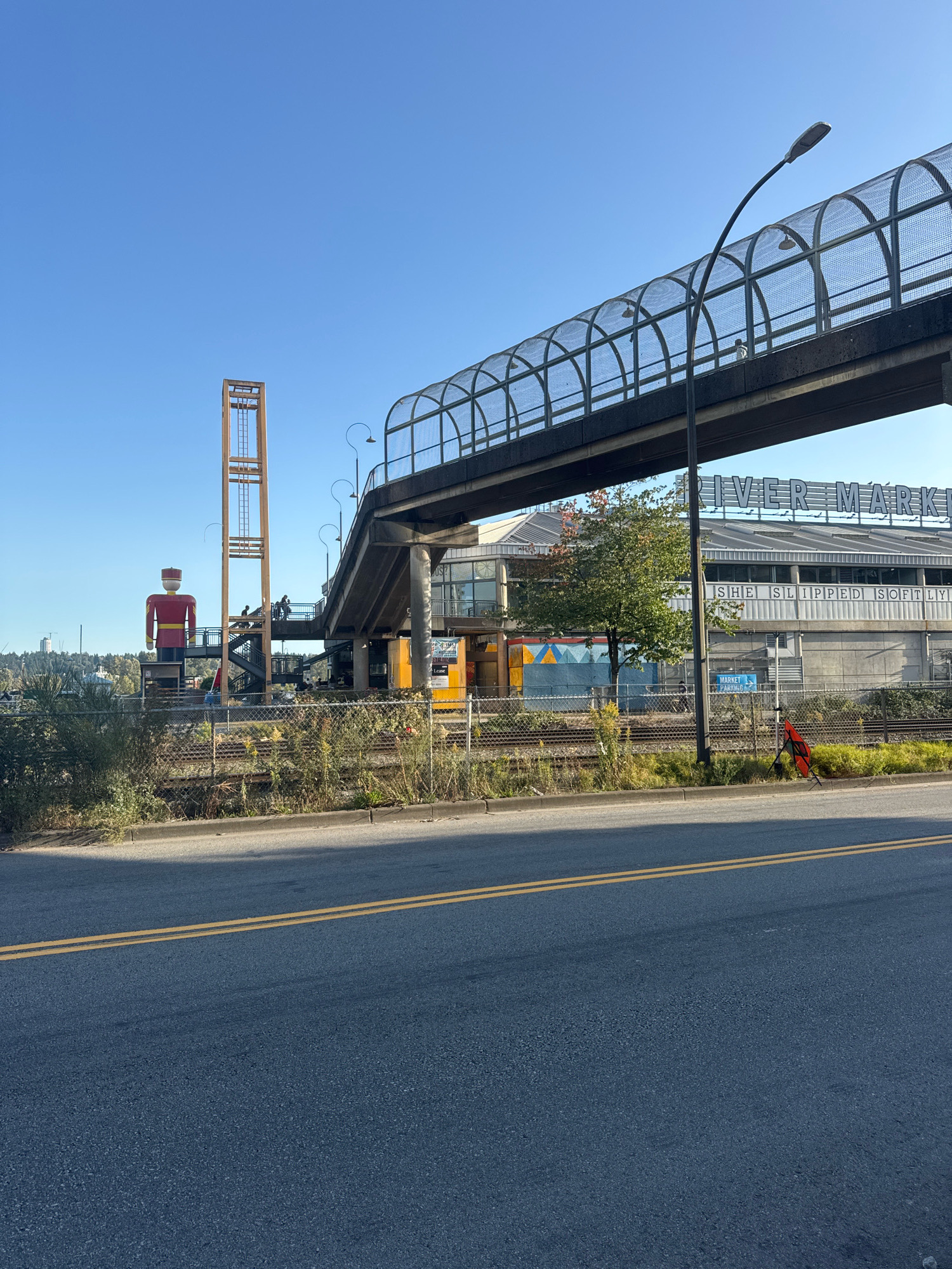 The image shows a street-level view of the River Market in New Westminster, Canada, on a clear, sunny day. In the foreground, a paved road with double yellow lines runs horizontally. Behind the road is a chain-link fence with dry grass and weeds growing through it.
Dominating the middle ground is a pedestrian overpass with a metal mesh enclosure. Below the overpass, train tracks are visible. To the left of the overpass, a tall, colorful, stylized figure resembling a soldier in a red and gold uniform stands beside a wooden tower structure. Several people are seen on the stairs and walkways of the overpass.
The River Market building itself is visible in the background, with large lettering spelling out "RIVER MARKET" across its roofline. Below this, the building features a grey facade with windows and a mural on a lower section. A sign indicating "MARKET PARKING" is also visible. A single tree with some green and reddish foliage stands near the market building.
The sky is a clear, bright blue. The overall scene depicts an urban environment with distinct landmarks and infrastructure.