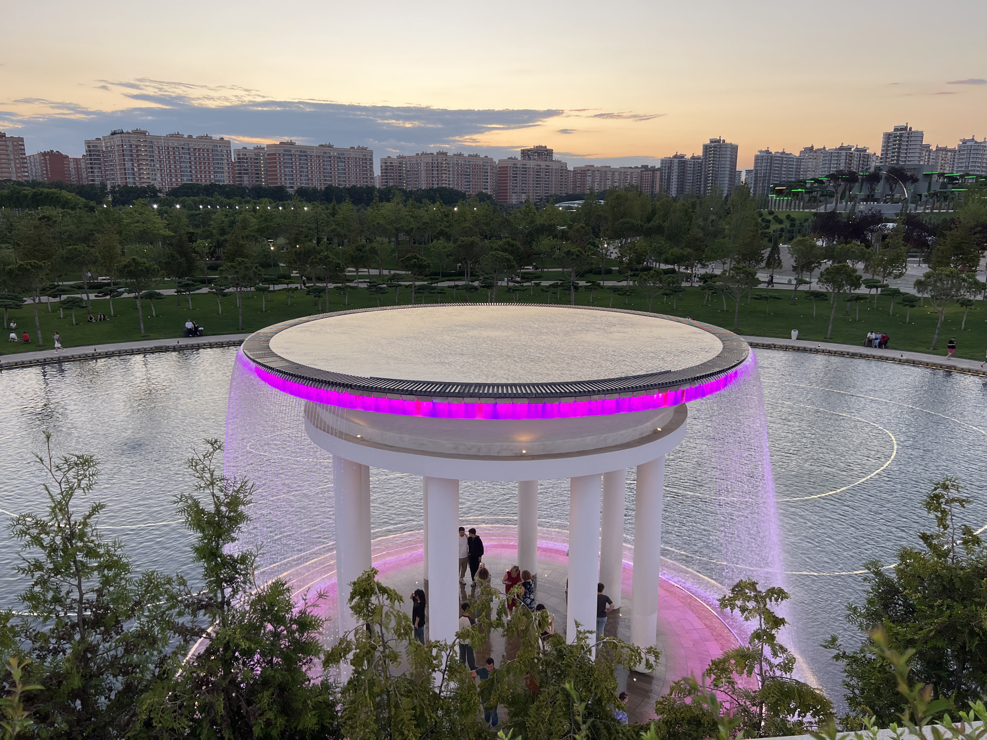 This media file captures an exterior view of a modern park with a prominent water feature at dusk. The central subject is a circular, open-air pavilion with white columns, topped by a flat roof. The roof is illuminated with vibrant pink lights along its edge, and water cascades down from it, creating a curtain effect. Several people are visible in and around the pavilion, some standing and conversing, while others appear to be observing the fountain.
The pavilion is situated within a large body of water, likely a pond or lake, with visible ripples and subtle lighting effects on the water's surface. In the background, a treelined park stretches out, with manicured lawns and scattered trees. Beyond the park, a dense cluster of modern apartment buildings forms the cityscape. The sky above is filled with soft clouds, transitioning from daylight to twilight, with hues of orange and purple indicating the time of day is close to sunset.
The overall scene suggests a public recreational space, possibly a park or garden, in an urban setting. The lighting, both the pink illumination of the fountain and the ambient twilight, creates a serene and visually appealing atmosphere. The presence of people enjoying the space points to a leisurely activity. No visible text is present in the image. The location is identified as Krasnodar, Russia.