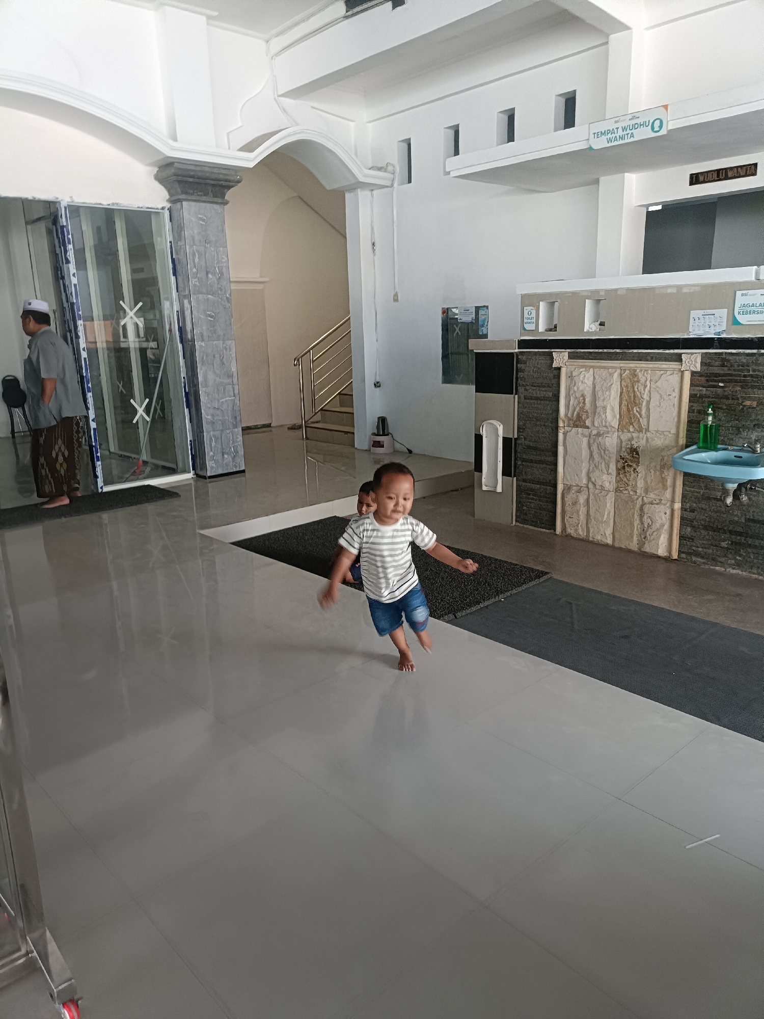 The image shows a young child running across a polished tile floor in what appears to be a public restroom area. The child is wearing a striped t-shirt and blue shorts. In the background, to the left, an adult male wearing a white cap and traditional Islamic attire is standing near a glass door. To the right, there is a sink with blue tiles and a soap dispenser. Signs are visible on the wall, one indicating "TEMPAT WUDHU WANITA" (Women's Ablution Area) and another in Indonesian stating "JAGALAH KEBERSIHAN" (Keep Clean). The environment suggests a clean and functional public facility, likely within a mosque or a similar religious building in Kediri City, Indonesia. The lighting is bright, indicating daytime.