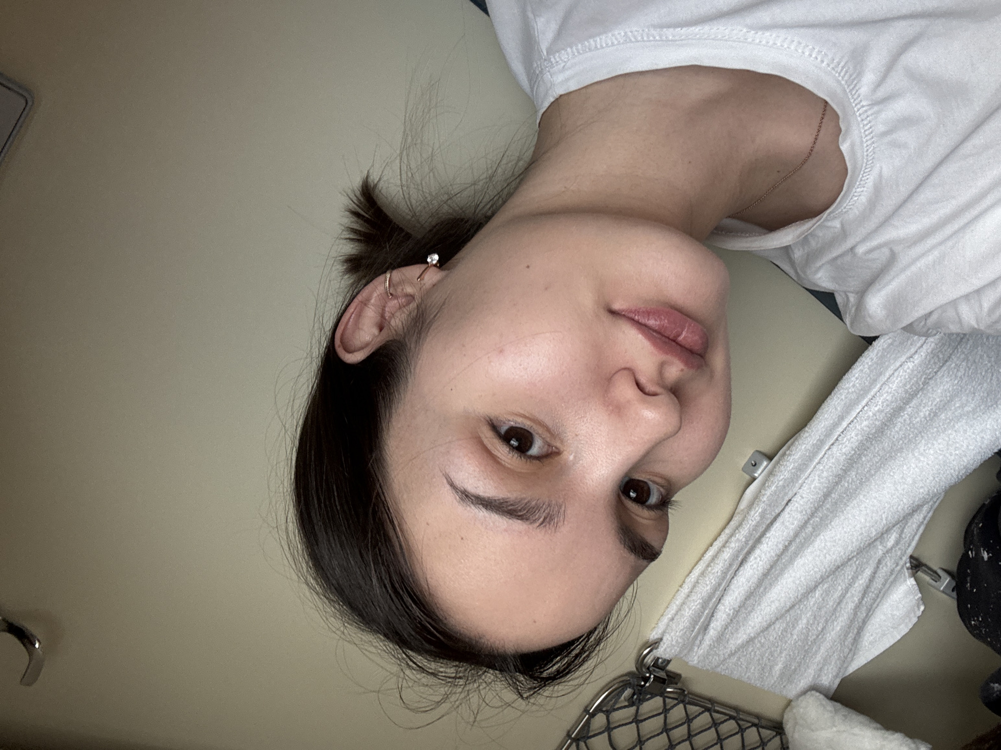 The image displays a close-up, angled selfie of a female individual inside what appears to be a train carriage. The subject, a young adult female with dark hair styled back, possesses light to medium skin tone and dark eyes that engage directly with the camera. Her expression is neutral with a slight upward turn of the lips. She wears a white crew-neck t-shirt and a thin, gold-colored chain necklace. Her visible right ear is adorned with a small, clear gemstone stud earring in the lobe and a gold-colored hoop or cuff in the upper cartilage.

The background features a light, neutral-colored wall, typical of an interior compartment. In the upper left quadrant, a curved metallic hook is affixed to the wall. Below this, a portion of a metal mesh net or basket, consistent with a storage rack, is visible, secured to the wall by metal clips. A white, textured towel is draped beneath the mesh, also attached to the wall with metal clips. A dark, indeterminate object is partially visible at the bottom left. The visual information does not provide details to ascertain a specific location within the city of Щучье Озеро, Russia.