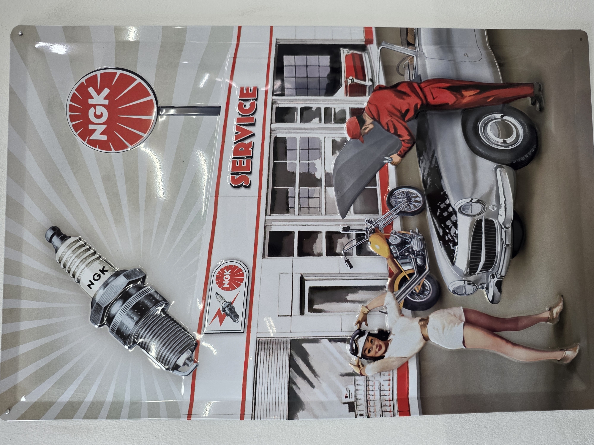 The image is a rectangular tin sign depicting an automotive service scene. The background is a light grey with radial stripes emanating from the left side. A white building labeled "SERVICE" with red trim and black-and-white windows is prominent. In the foreground, there's a chrome spark plug with "NGK" printed on it. A red and white NGK logo is present on the sign and near the building.
Overlapping the building and background are images of a grey vintage car, a yellow motorcycle, a mechanic in a red uniform working under the car's hood, and a woman in a white outfit lying down near the motorcycle. The overall impression is a collage-style automotive advertisement. There is no visible evidence to link this image to Nicosia, Cyprus.