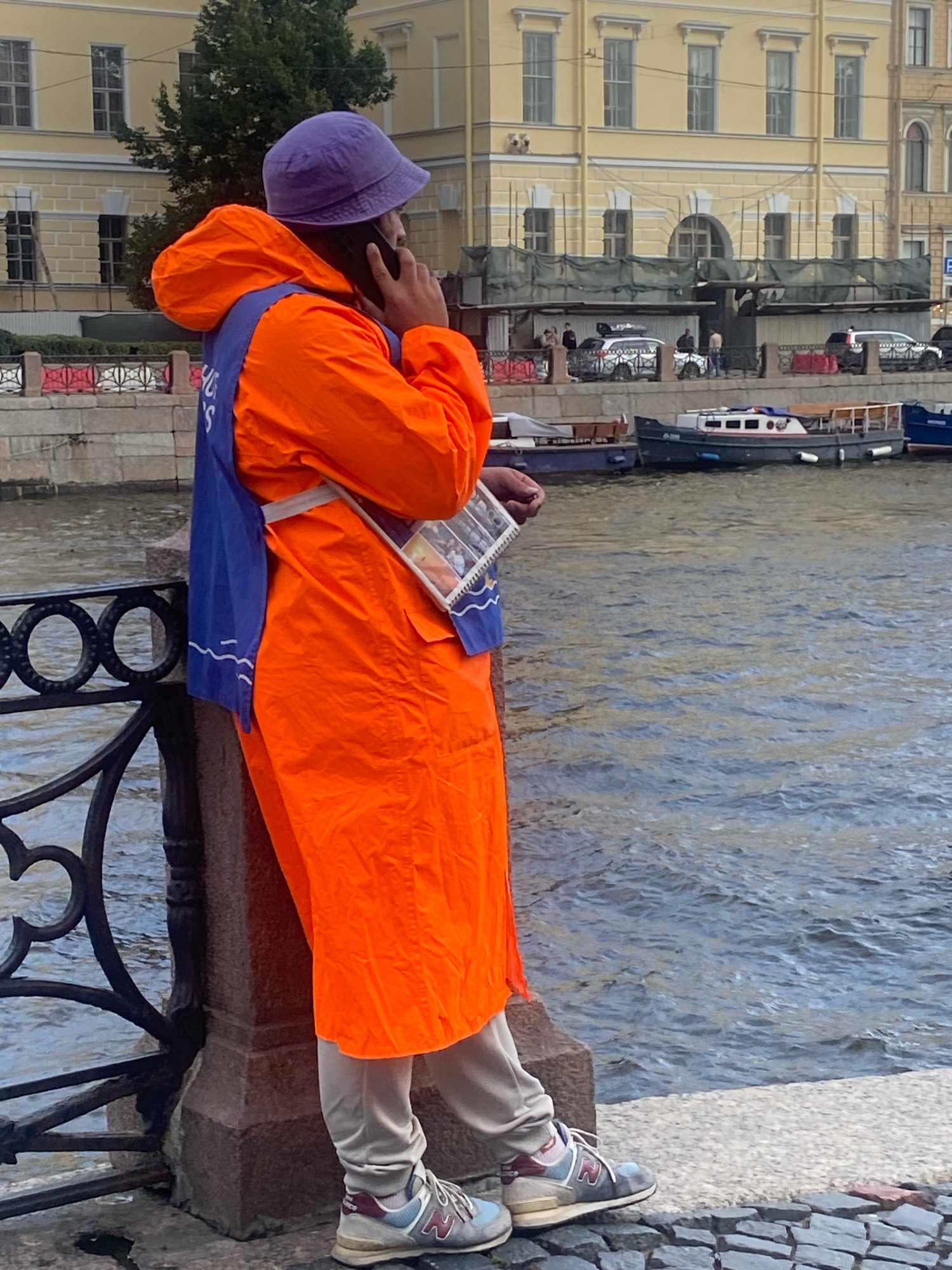 The image shows a person wearing an orange hooded jacket and a purple bucket hat, standing near a body of water in Saint Petersburg, Russia. The person is holding a phone to their ear and has a blue vest over the orange jacket. They are also holding a rectangular object with images on it. In the background, there is a building with yellow walls and arched windows, and several boats are docked on the water. The person is standing on a paved walkway next to a decorative metal railing. The overall impression is of a person on a phone call in a public space, likely a tourist area, given the presence of boats and the architecture.
