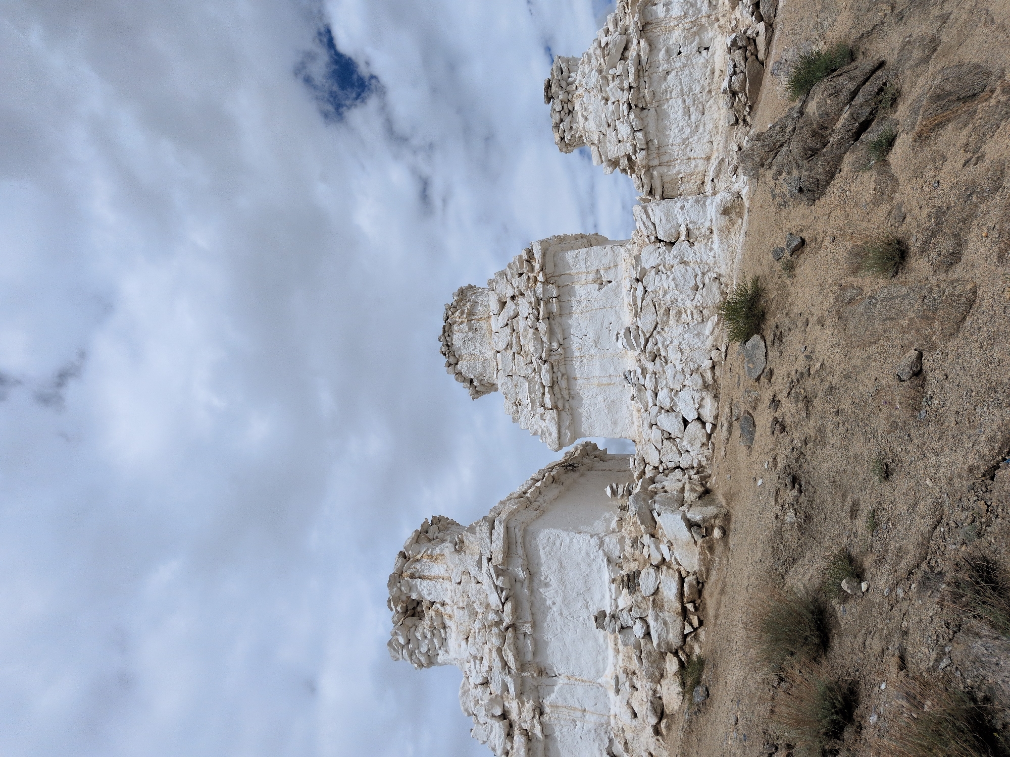 Three white stupas are situated on a hillside under a cloudy sky. The stupas are built of stone and plastered white, with rounded tops. They are of varying sizes, with the largest one in the center, and two smaller ones flanking it. The hillside is covered with dry earth, rocks, and sparse vegetation. The stupas appear ancient and weathered, with some damage to their structures. The overall scene suggests a remote and possibly religious site. The stupas are located in the city of Leh, India.