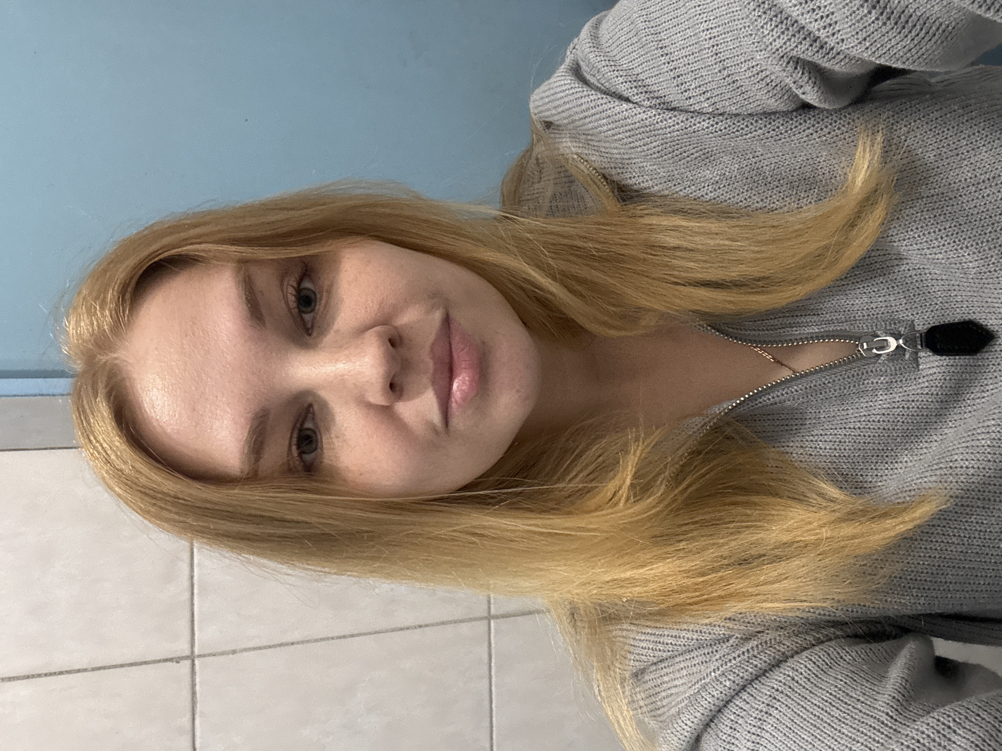 A fair-skinned woman with long blonde hair and light-colored eyes is centered in the image. She wears a gray knitted zip-up sweater. The background consists of a blue painted wall in the upper part of the image and white tiled wall in the lower part of the image. The image is taken indoors and rotated sideways. Rusanovo, Russia, is not apparent from the image.
