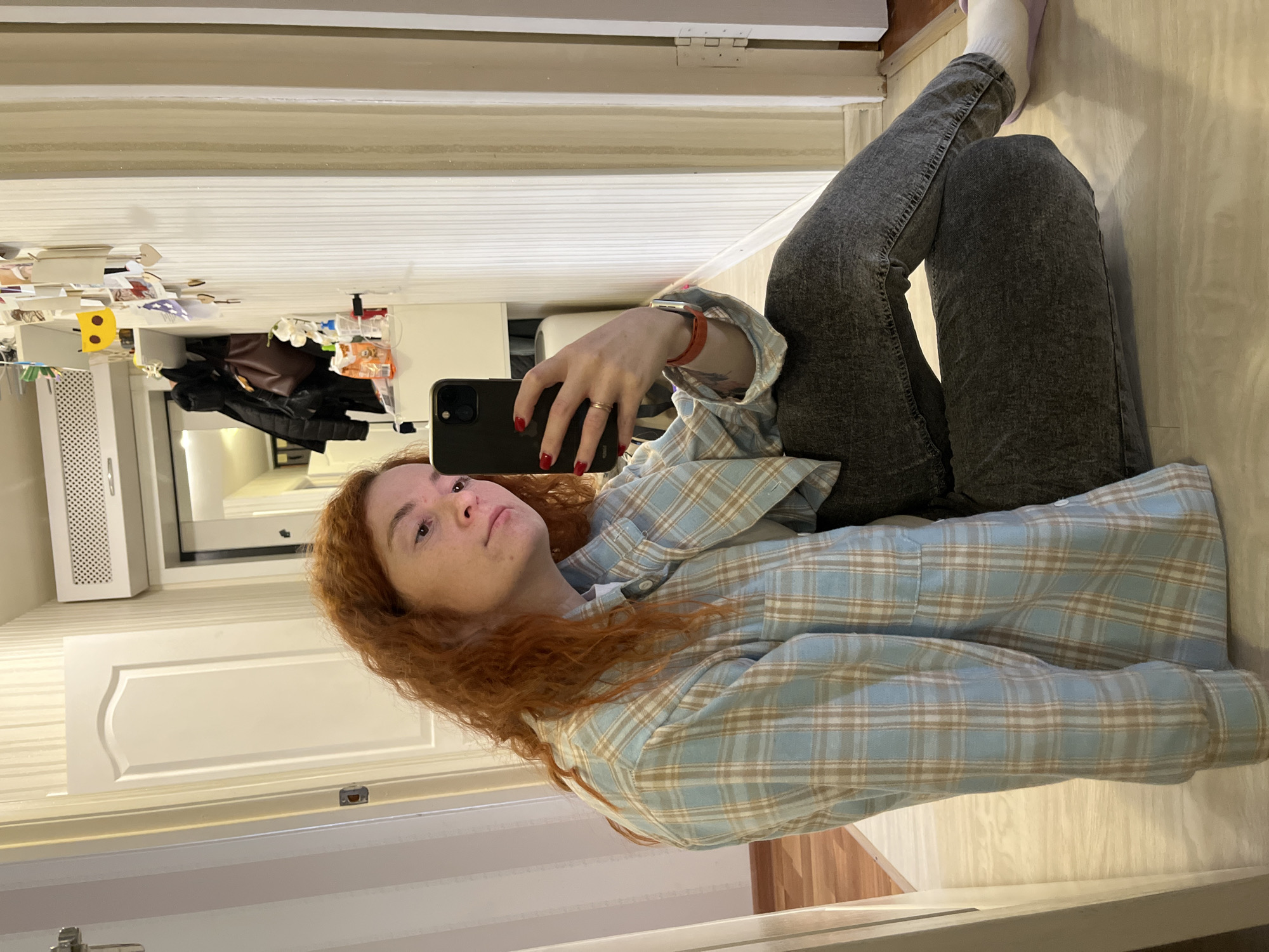 A woman with red hair is seen in an indoor setting, likely a home in the city of Chaikovsky, Russia. She is wearing a light blue and tan plaid shirt and dark grey jeans, with a white sock visible on her right foot. She is holding a black smartphone, presumably taking a selfie. The background includes a hallway with a white door, white walls, and a white cabinet with items on top. A mirror reflects the hallway, showing clothing items hanging inside a closet. The floor is a light beige color.