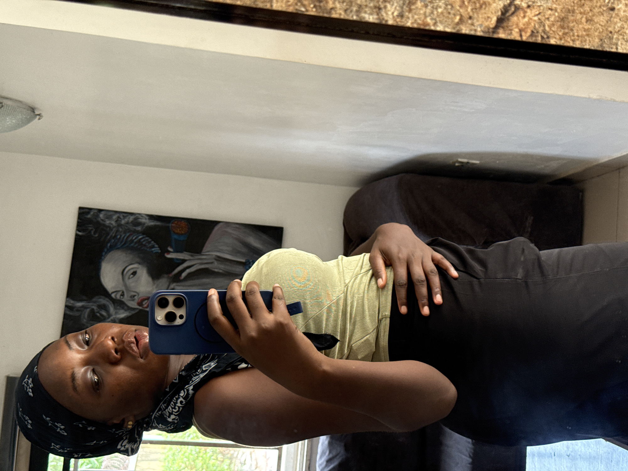 A woman is taking a selfie in a mirror. She is wearing a black headscarf and a light-colored top, and black pants, holding a blue smartphone. Behind her is a painting on the wall, a couch, and a window. The location is likely an apartment in Abuja, Nigeria, based on the interior style and the woman's appearance.