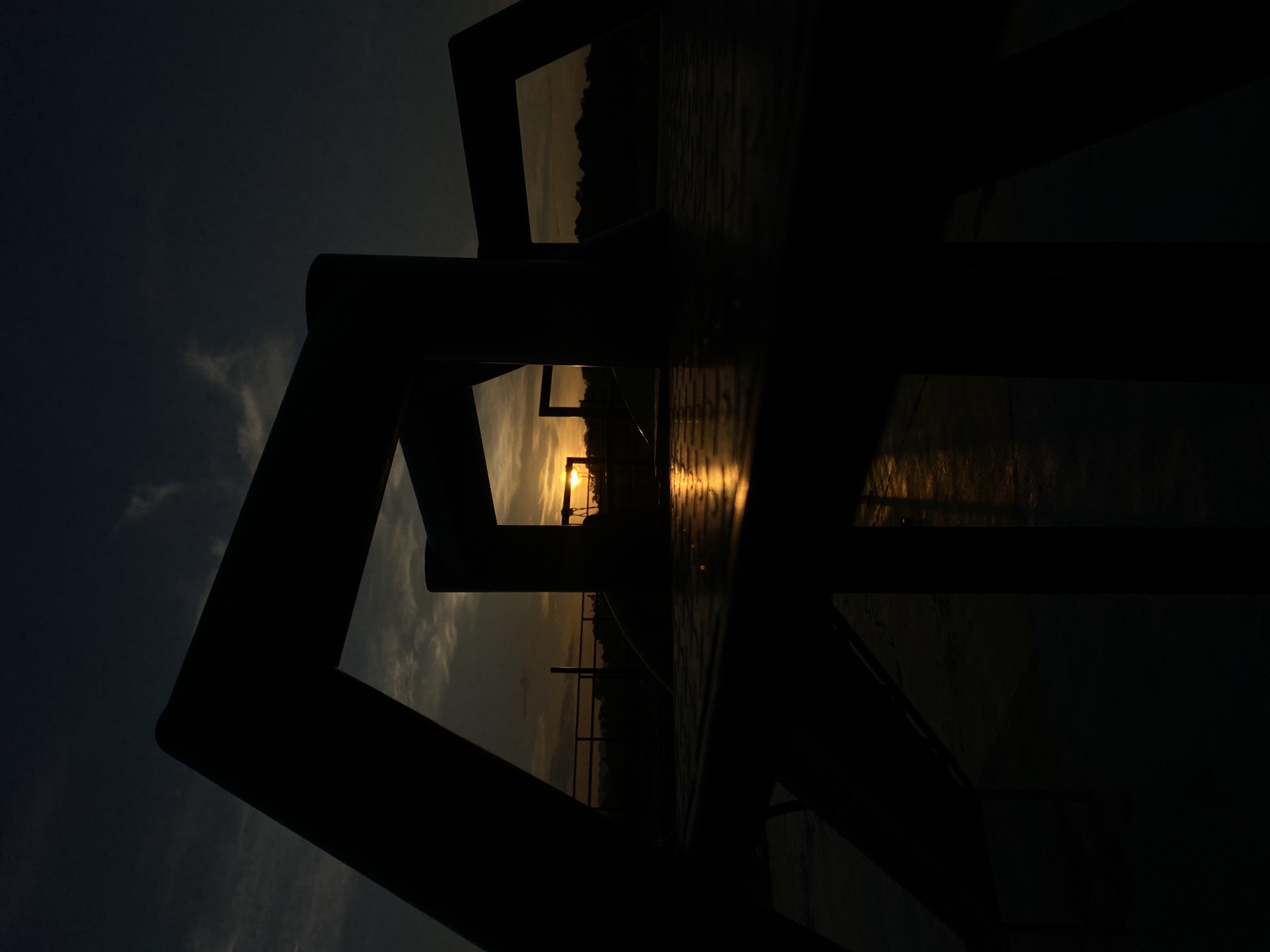 The image captures a sunset viewed through the silhouette of playground equipment. The sun is low on the horizon, casting a warm glow and reflecting off a wet surface, possibly a metal deck or slide. The foreground is dominated by the dark, geometric shapes of the playground structure, creating a strong contrast against the sky. In the background, a treeline is visible, silhouetted against the fading light, with a railing running horizontally across the middle ground. The sky is a gradient of dark blues and greys, with wisps of clouds catching the last rays of the sun. This scene is situated in an outdoor public space, likely a park or playground, common in urban areas such as Davao City, Philippines, where recreational facilities are often found. There are no people or animals visible in the frame.