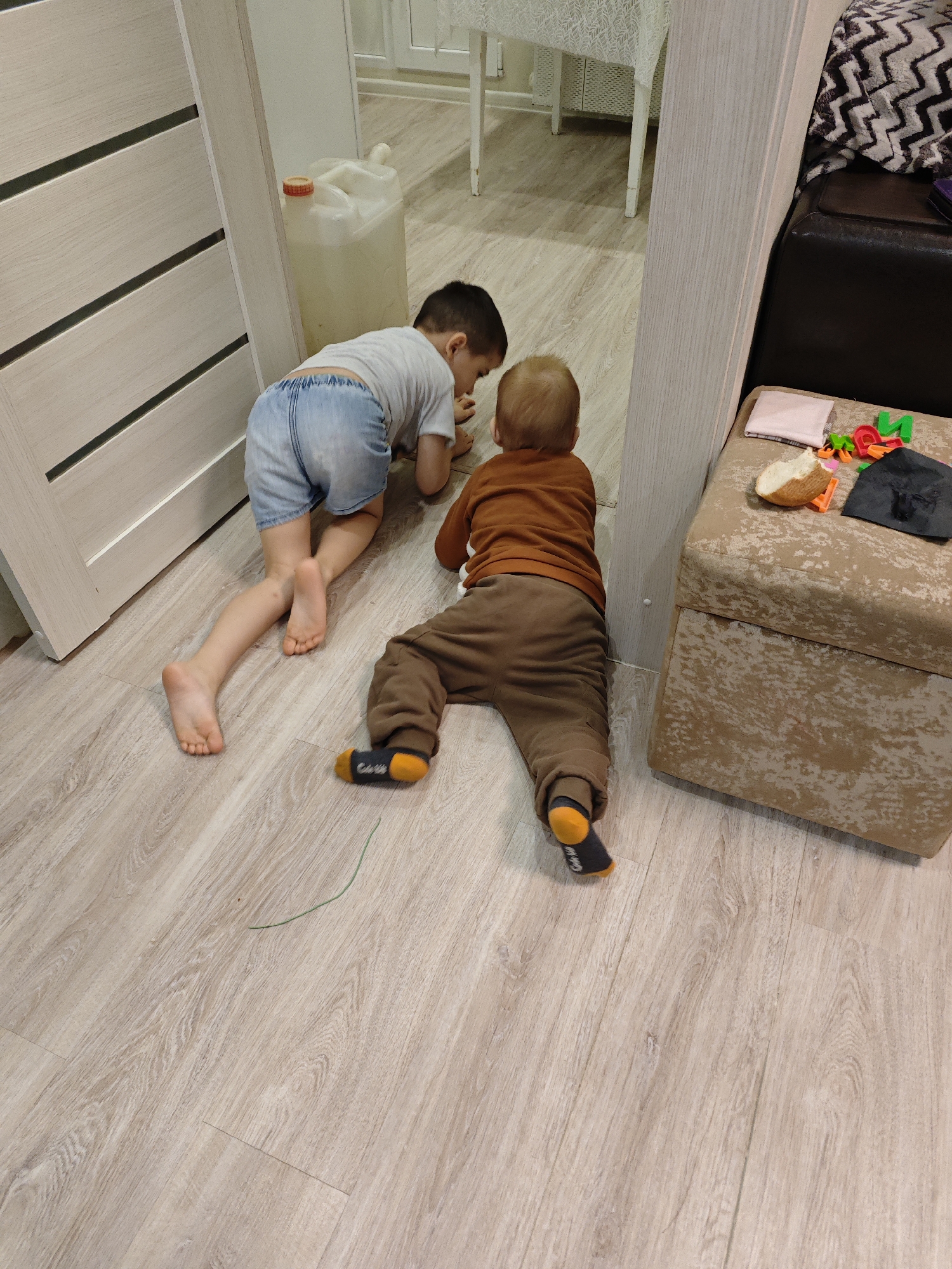 Two children are on a wood-look floor. A fair-skinned child in denim shorts and a gray t-shirt is kneeling, facing towards the left. Another child, also fair-skinned, wearing brown pants and an orange long-sleeved shirt, is crawling. The scene appears to be indoors, in a room with a light-colored door on the left and a beige ottoman with a slice of bread, some alphabet magnets, and a black cloth on the right. A plastic water container sits between the children and a white table. The location is Chaikovsky, Russia.