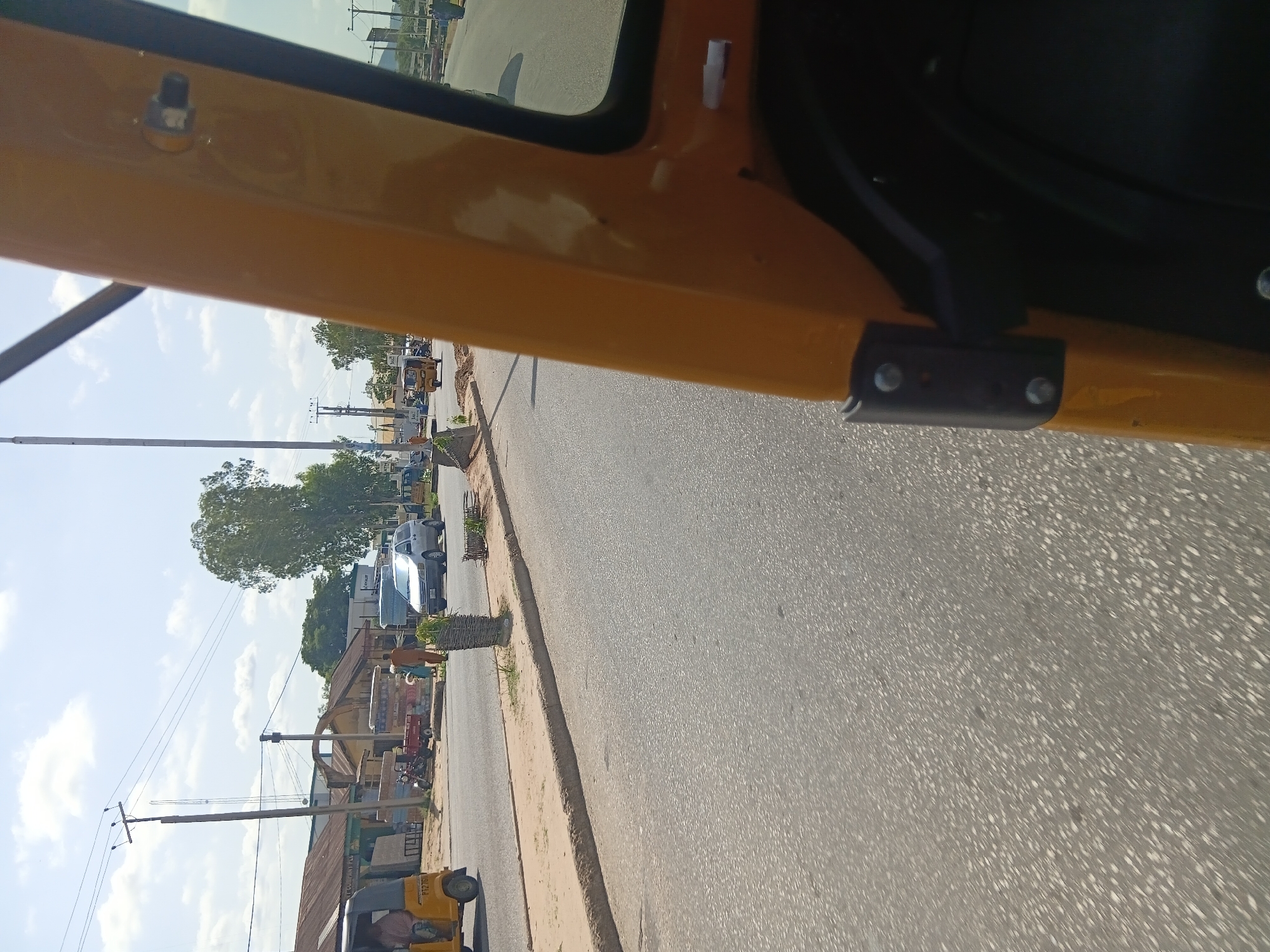 The image captures a street scene in Jalingo, Nigeria, likely taken from within a yellow auto-rickshaw. The foreground is dominated by the yellow body of the auto-rickshaw, showing a portion of the passenger compartment and a black interior. A black metal bracket with silver bolts is attached to the yellow body. The road is paved with asphalt, exhibiting a speckled texture. To the left of the road, a sandy shoulder leads to a paved sidewalk.
Along the sidewalk, a few vehicles are visible. A yellow auto-rickshaw with license plate "P12701" is parked on the far left. Further down, a red motorcycle with a small trailer is parked next to a small building with a corrugated metal roof. A silver van or SUV is driving on the road, and another auto-rickshaw is visible further in the distance. People are seen walking on the sidewalk and near the parked vehicles.
The background features a row of buildings with corrugated metal roofs, typical of commercial establishments. A tall tree with lush green foliage stands prominently in the middle ground. Beyond the buildings and trees, utility poles with overhead wires are scattered across the landscape, indicating the presence of infrastructure. The sky is blue with scattered white clouds, suggesting a clear, sunny day. The overall environment depicts a bustling urban street in Jalingo.