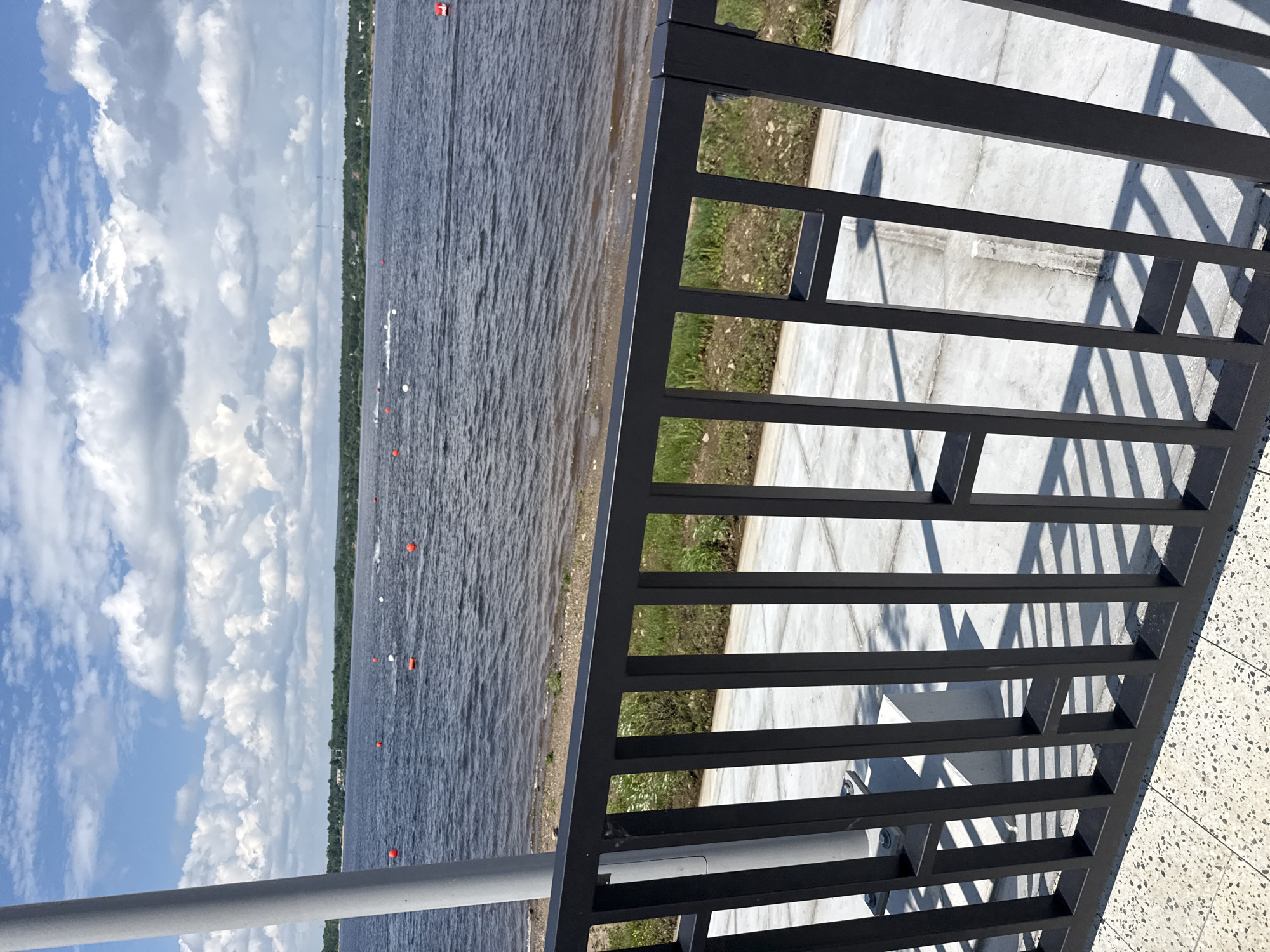 The image captures a wide shot of a body of water, likely a river or lake, under a partly cloudy blue sky. In the foreground, a black metal railing with vertical bars and horizontal rectangular openings borders a paved area. Beyond the railing, a strip of gravel and grass leads to the water's edge. The water itself is dark and choppy, dotted with several orange and white buoys. In the distance, a tree-lined shore is visible with a few structures and distant power lines. Some individuals or objects are moving on the water, indicated by white wakes. The scene appears to be a public waterfront area, possibly a park or promenade. Based on visual cues, the location is consistent with the Kama River embankment in the city of Perm, Russia, particularly areas like the Permskaya embankment or waterfront parks. The presence of buoys suggests a possible water sports event or marked swimming area.