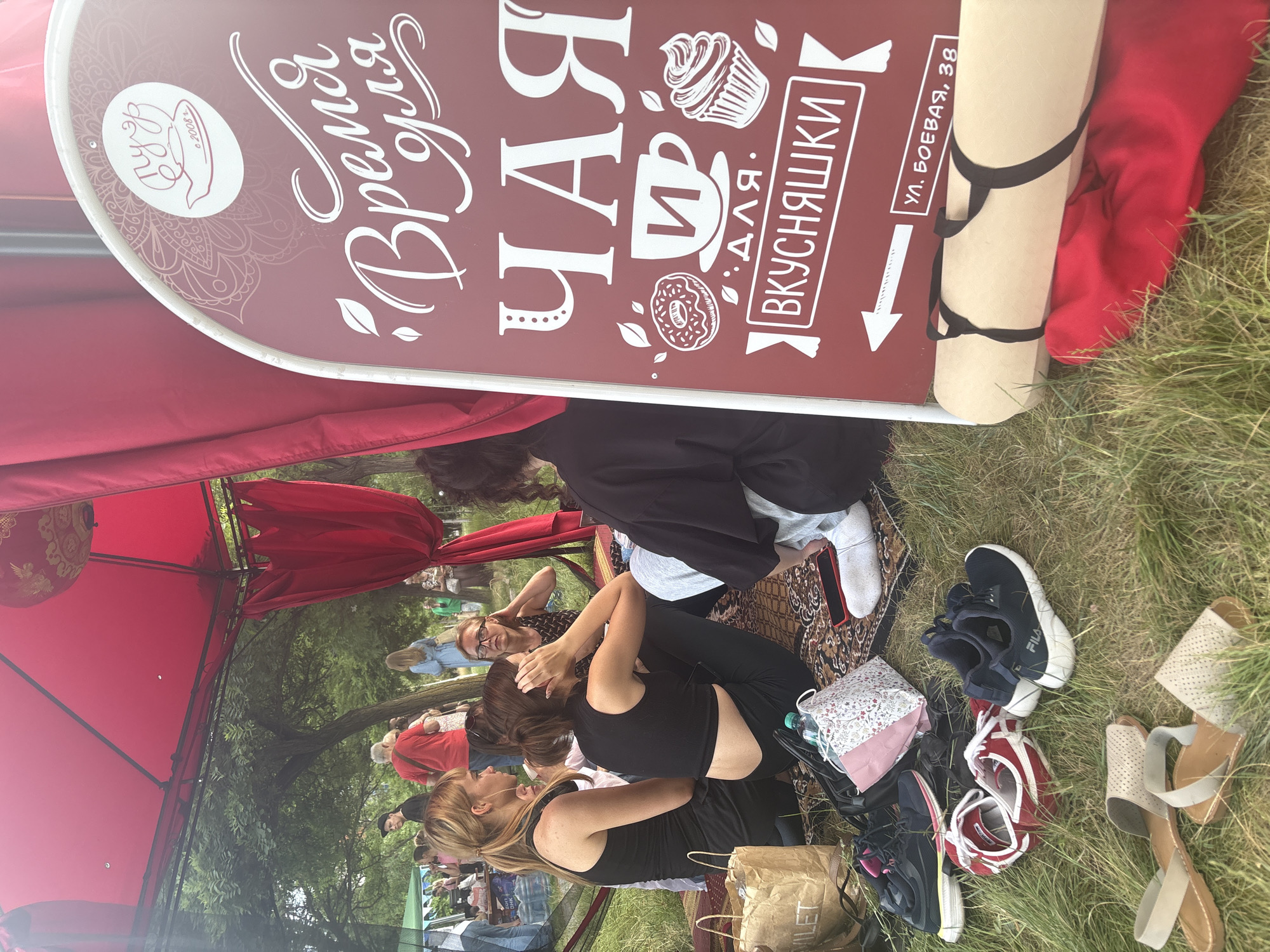 A group of people are gathered on a rug under a red canopy. A burgundy sign with white text and illustrations is propped up to the right of the group. The sign reads "Время для чая и вкусняшки" (Time for tea and treats) and provides an address "ул. Боевая, 38" (Boevaya St, 38). Several pairs of shoes are scattered on the grass in front of the group. The environment appears to be a park or outdoor gathering space with trees visible in the background.