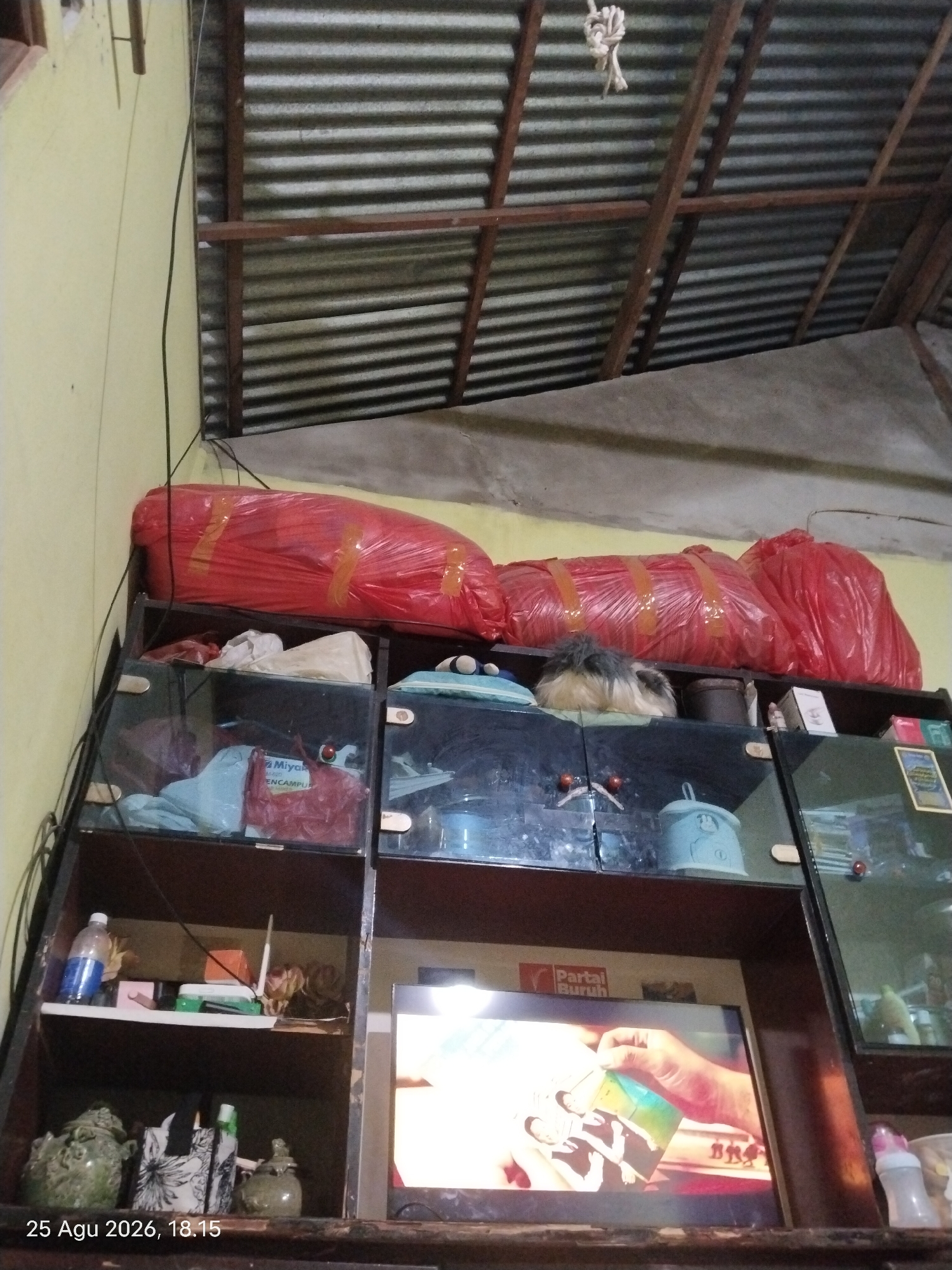 The image depicts an indoor scene, possibly a storage area or a room in a residence, located in Singkawang, Indonesia, as indicated by the timestamp "25 Agu 2026, 18.15". The overhead structure features corrugated metal roofing supported by wooden beams. Below this, a shelf unit is visible. On the top shelf, three large, red plastic-wrapped items are stacked horizontally. These items appear to be bags or bundles, secured with yellow tape. Below this, the shelf unit has glass-fronted compartments containing various objects. In one compartment, there are several items wrapped in plastic, including a blue bag and a red bag with text that appears to be "Miyaka" and "ENCAMPUR". Another compartment contains a light blue cylindrical object. On a lower shelf, a clear plastic bottle containing a blue liquid is present, along with other items such as a small orange box, a white antenna, and dried floral arrangements. The bottom section of the shelf unit houses a television screen displaying an image of a hand holding a booklet with people on it, and a logo that reads "Partai Buruh". To the left of the television are decorative green ceramic jars and patterned bags. To the right of the television, a bottle with a pink nipple is visible, suggesting it is a baby bottle. The overall environment appears cluttered and utilitarian.