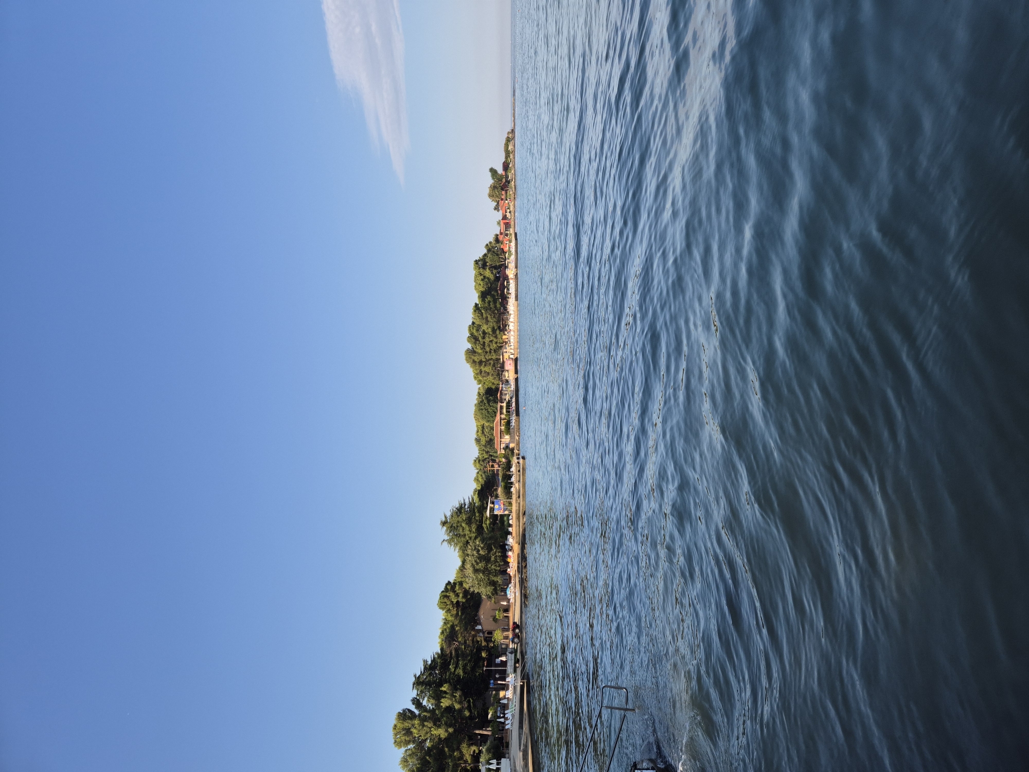 The image shows a tranquil coastal scene in Monterol, Croatia. The foreground is dominated by the deep blue, rippling surface of the Adriatic Sea. In the middle ground, a sandy beach is visible, lined with lush green trees. Scattered across the beach are lounge chairs and umbrellas, suggesting a resort or recreational area. Several small buildings with red roofs are nestled among the trees, appearing to be part of the resort. The sky is a clear, vibrant blue with a few wispy white clouds on the horizon. To the left of the image, a set of metal stairs leads down into the water from a paved walkway, hinting at a place where people might enter or exit the sea. The overall atmosphere is peaceful and idyllic, indicative of a sunny day at a seaside location.