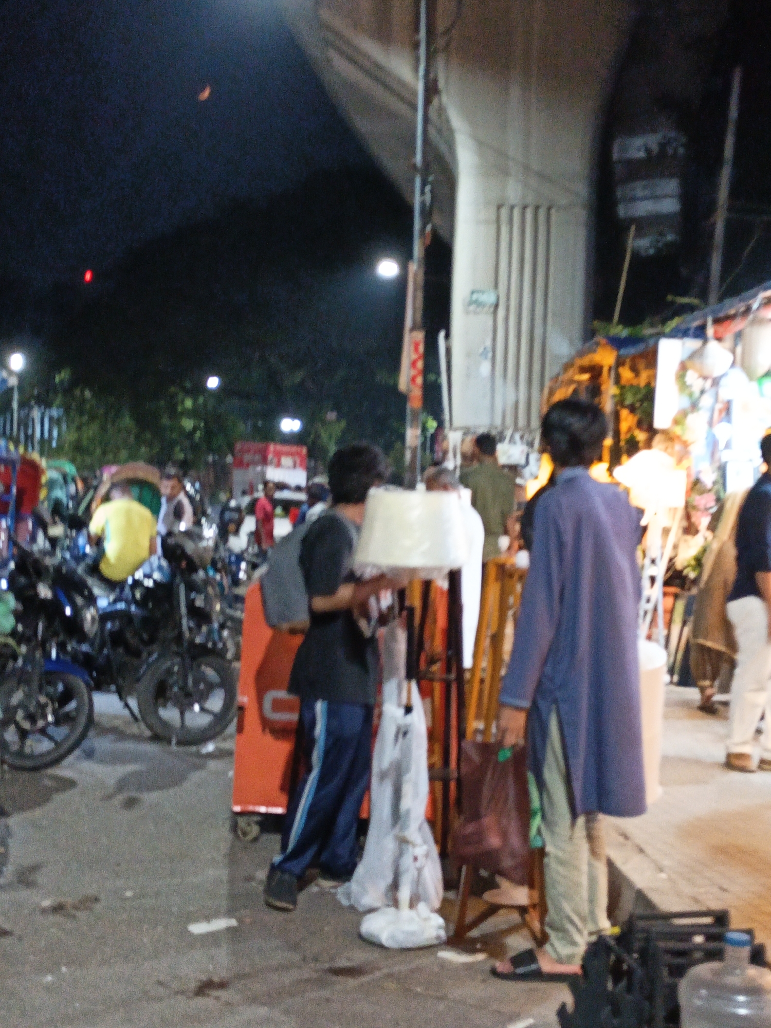 An outdoor, night scene in an urban environment, possibly a marketplace, is depicted. A bridge or overpass looms in the background. In the foreground, motorcycles are parked. Several people are visible. One person, in the center of the frame, is wearing a dark shirt and blue pants with white stripes and is holding a white object. To the left, another person is wearing a blue tunic and light-colored pants, holding a dark plastic bag. To the right, a person in a long blue garment is visible from behind, and further back, a person in white pants is seen. The ground appears to be asphalt.