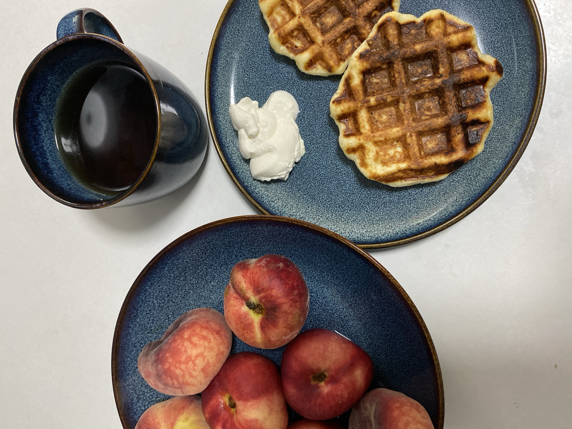 This overhead shot shows a breakfast spread on a white surface. In the upper left is a dark blue, speckled mug filled with black coffee. To its right, a matching blue plate holds two golden-brown waffles and a dollop of white cream. Below this, another blue plate is filled with several flat, round peaches, showing their reddish-pink skin and yellow flesh. The scene appears to be set indoors, likely in a home or cafe, given the casual presentation of the food and drink. The lighting is bright and diffused, suggesting daytime. The overall mood is peaceful and suggests a relaxed morning meal. There is no visible text in the image.