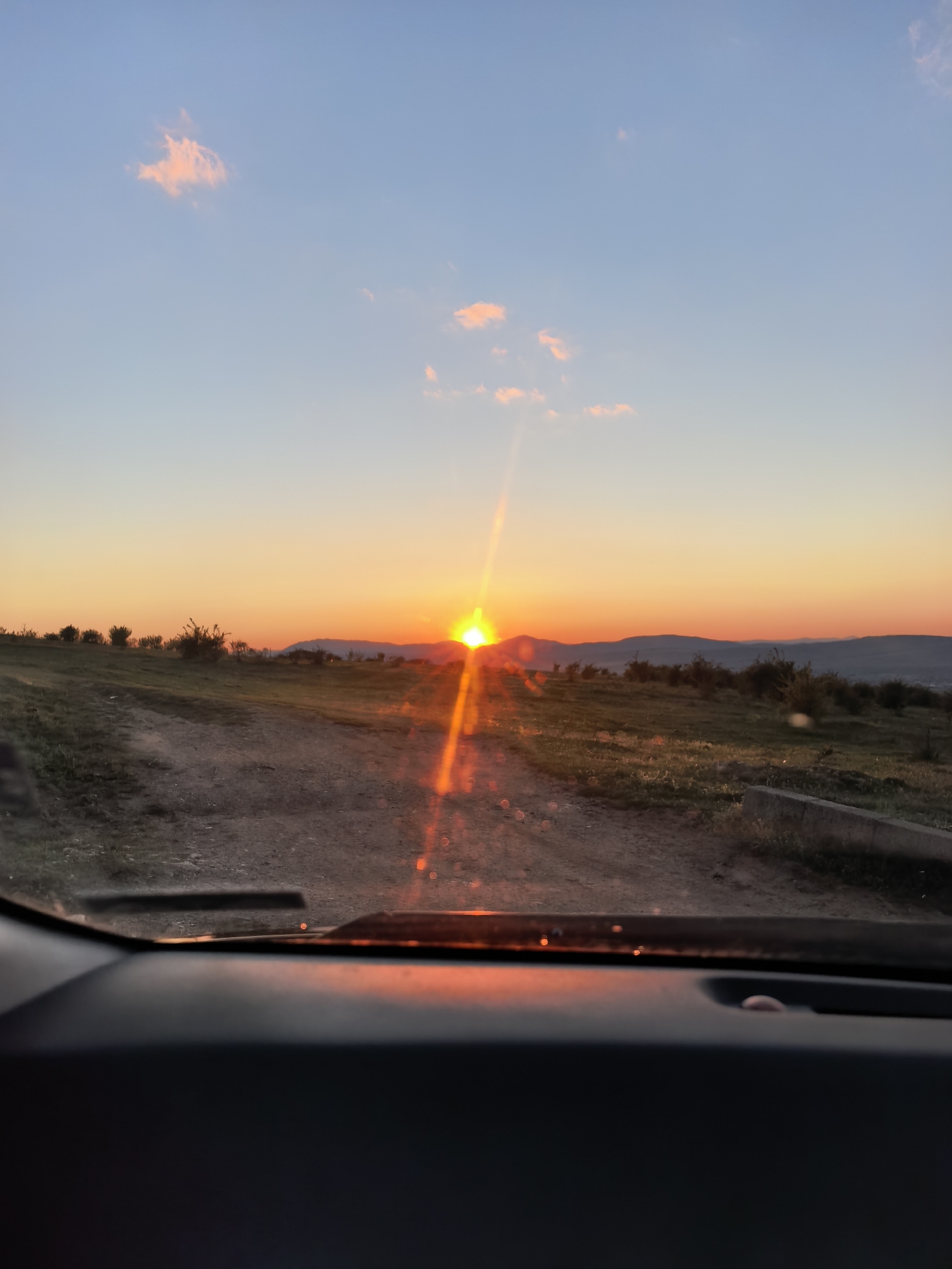 The image depicts a sunset over a rural landscape, as viewed from inside a vehicle. The sun is setting directly ahead, casting a bright orange glow across the horizon and a light glare in the image. The sky is a gradient of blue to orange. A dirt road stretches towards the horizon, flanked by grassy fields and sparse vegetation. The dashboard of the vehicle is visible in the foreground. The landscape appears to be mountainous. This location is Sapareva Banya, Bulgaria.