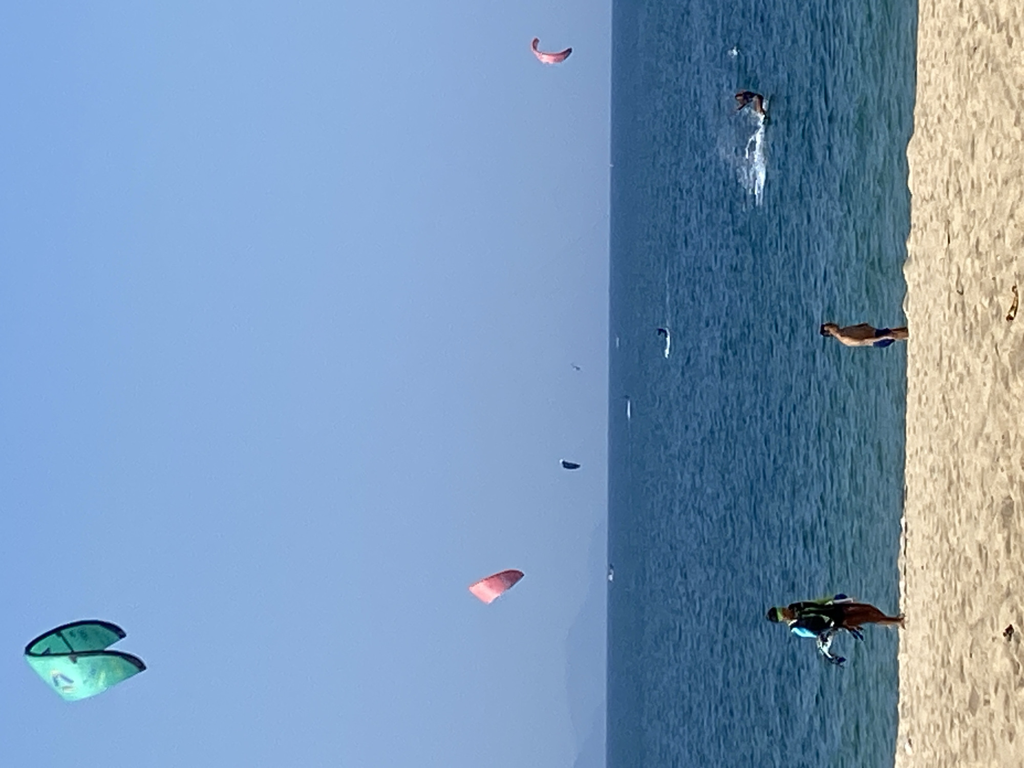 The image depicts a bright, sunny day at a beach, likely in Nha Trang, Vietnam. The scene is dominated by the vast expanse of the ocean under a clear blue sky. In the foreground, a sandy beach is visible.
Several individuals are engaged in watersports. In the middle distance, a person is kitesurfing, leaving a spray of water behind them. Further out in the ocean, there are at least two other kitesurfers or individuals engaged in similar activities. Various kites, in colors like green and pink, are visible in the sky, indicating active kitesurfing.
On the beach, a person is standing near the water's edge, holding a kite. To their right, a man is standing on the sand, looking towards the ocean. The water appears calm with gentle waves. The horizon line is defined by distant mountains or landforms. The overall impression is one of a recreational beach scene on a clear day.