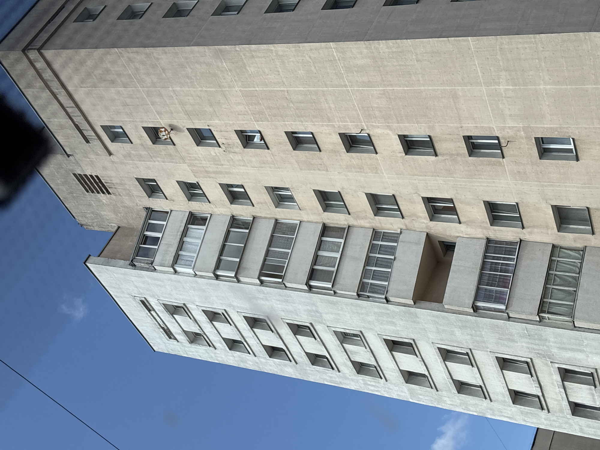 A tall, multi-story concrete apartment building dominates the frame, seen from a low-angle perspective against a clear blue sky with sparse clouds. The building features a repetitive pattern of windows and balconies. On the right side of the building, a single window on the sixth floor from the top displays a circular object, possibly a basketball hoop with a deflated ball, attached to the exterior. The building's facade is weathered and shows signs of age. A dark power line stretches diagonally across the upper left portion of the sky. This apartment building is located in Minsk, Belarus.