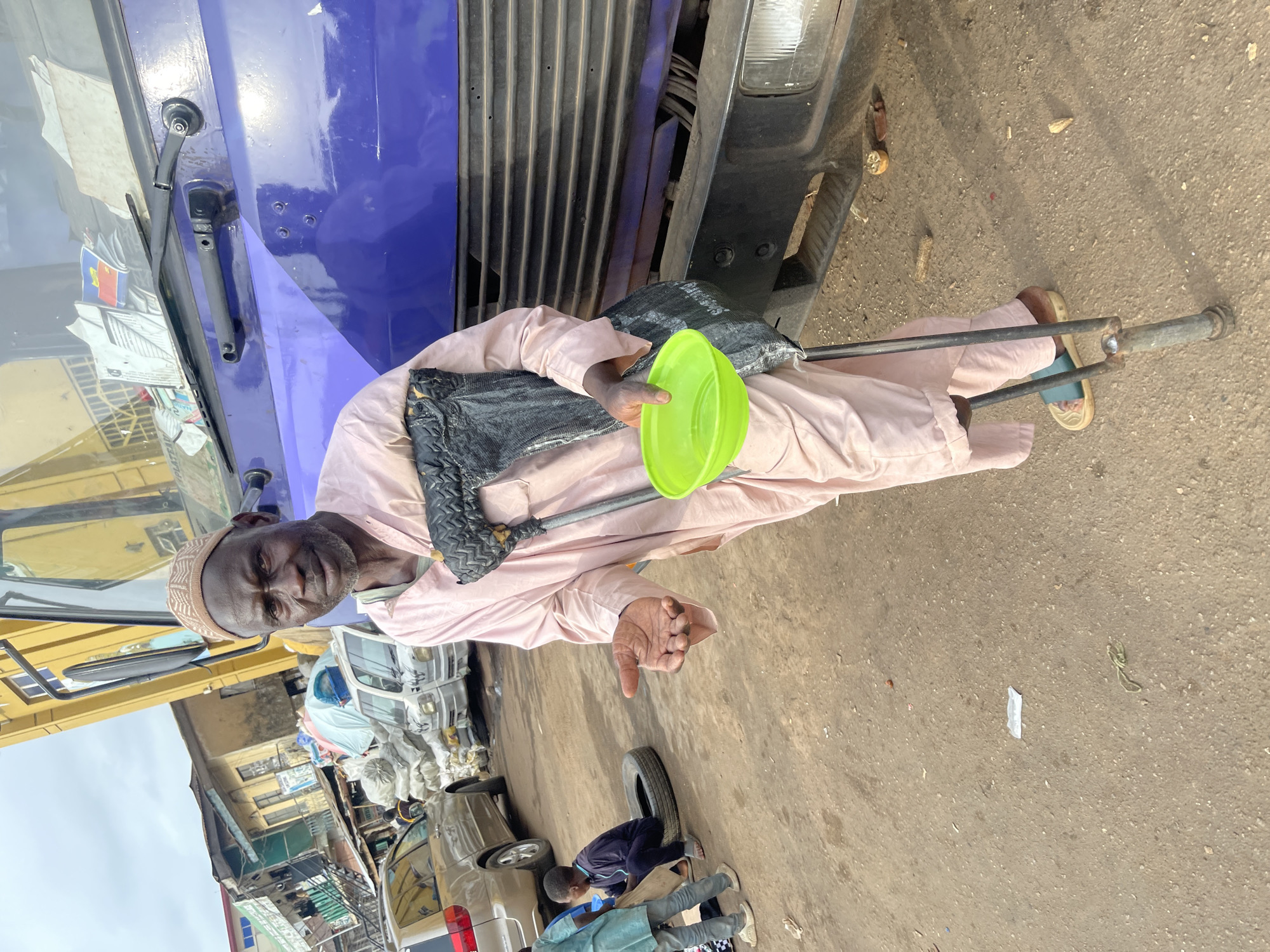 A man is standing in front of a blue and yellow truck in what appears to be a market area in Jos, Nigeria. He is wearing a light pink robe and a beige cap. He is using crutches for support and holding a bright green bowl in his outstretched hand, seemingly asking for donations. The ground is a mix of dirt and gravel, with some debris scattered around. In the background, there are several other vehicles, including a gold minivan, and buildings that suggest a commercial or residential area. The overall atmosphere appears to be that of a busy street market.