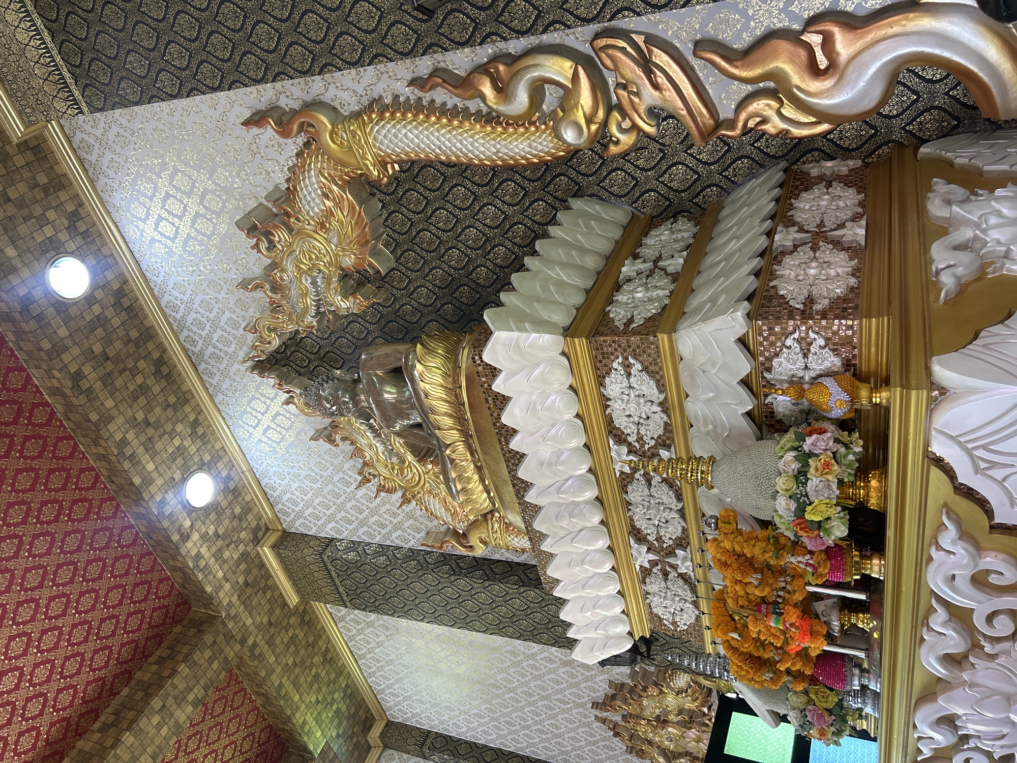 An ornate, multi-tiered altar dedicated to a Buddha statue is depicted in this image. The altar is adorned with golden dragon motifs and floral arrangements. The background features patterned walls and a gold-tiled ceiling with red accents. The overall impression is one of opulence and religious reverence. The location is the Wat Phra Non monastery in Thai Ko Subdistrict, Thailand.