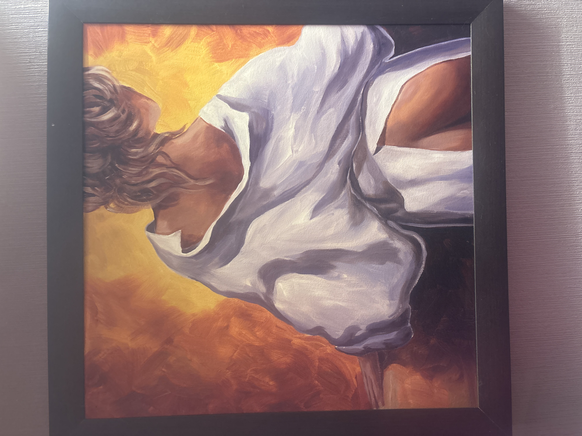 This media file features a framed painting, photographed as it hangs on a textured indoor wall. The painting is encased in a simple, flat black frame. The artwork itself is an expressive piece, likely an oil or acrylic painting, depicting the upper body of a person, presumably a woman. She is shown in a dynamic pose, bending backward or arching her neck to look upwards, with her head and wavy, flowing brown hair positioned towards the top left of the canvas. She is dressed in a loose, white garment, possibly a shirt or dress, which drapes fluidly over her shoulders and torso, revealing a portion of her chest.
The background of the painting is a vibrant array of warm hues, transitioning from a bright yellow at the top and left to deeper oranges and reddish-browns across the rest of the canvas. Visible brushstrokes contribute to a painterly style, suggesting movement and energy. The overall composition evokes feelings of freedom, passion, or intense emotion. The wall visible outside the frame features subtle horizontal ridges, indicating an indoor setting. There is no visible text in the image. This media file was captured in Ruse, Bulgaria.