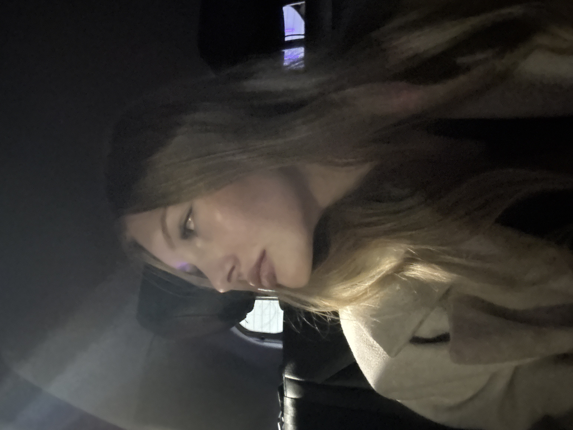 A young woman with long blonde hair is seen from the side, looking to her left. She is wearing a light-colored coat with a fur-lined collar. The setting appears to be the interior of a vehicle, with a dark headrest visible behind her. The background is dark, with some indistinct lights suggesting it is nighttime and potentially in an urban environment. The image does not provide any specific visual cues to determine a location within the city of Perm, Russia.