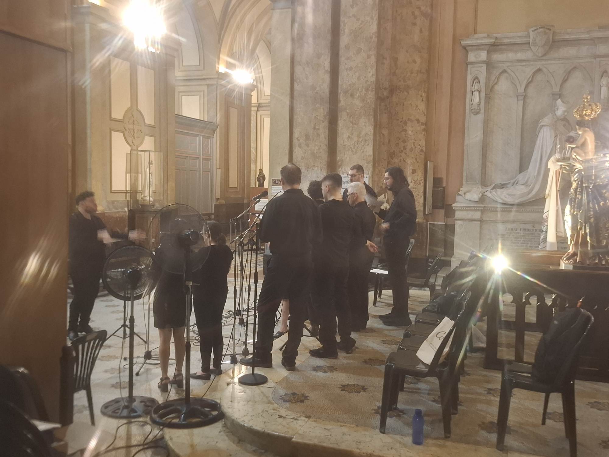 The image shows a group of people in what appears to be a church in Buenos Aires, Argentina. The men and women are dressed mostly in black, and some appear to be setting up microphones. There are folding chairs in rows. The location is suggested by the architecture and the presence of a monument with the inscription "LEONE FRIDERICO".