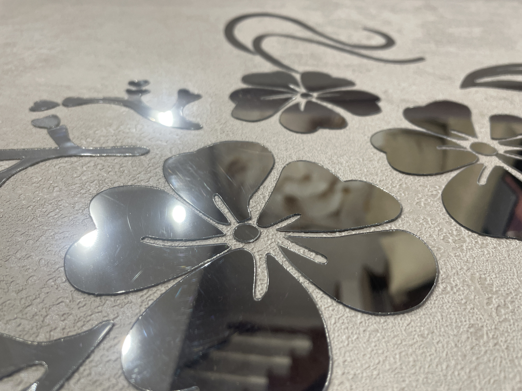 The image shows a close-up view of decorative mirrored floral decals affixed to a textured, light-colored wall. The decals are silver and reflect the surrounding environment, including what appear to be furniture and possibly lights. The floral designs are stylized, with distinct petal shapes and central circular elements. The texture of the wall is rough, resembling a stucco or heavily textured wallpaper. The lighting in the scene causes bright highlights and some glare on the mirrored surfaces, obscuring some details. There are no people or explicit actions depicted. The location is noted as Aropakkuzi, Russia, though the image itself does not provide visual cues to confirm this specific geographical context beyond what is provided in the prompt.